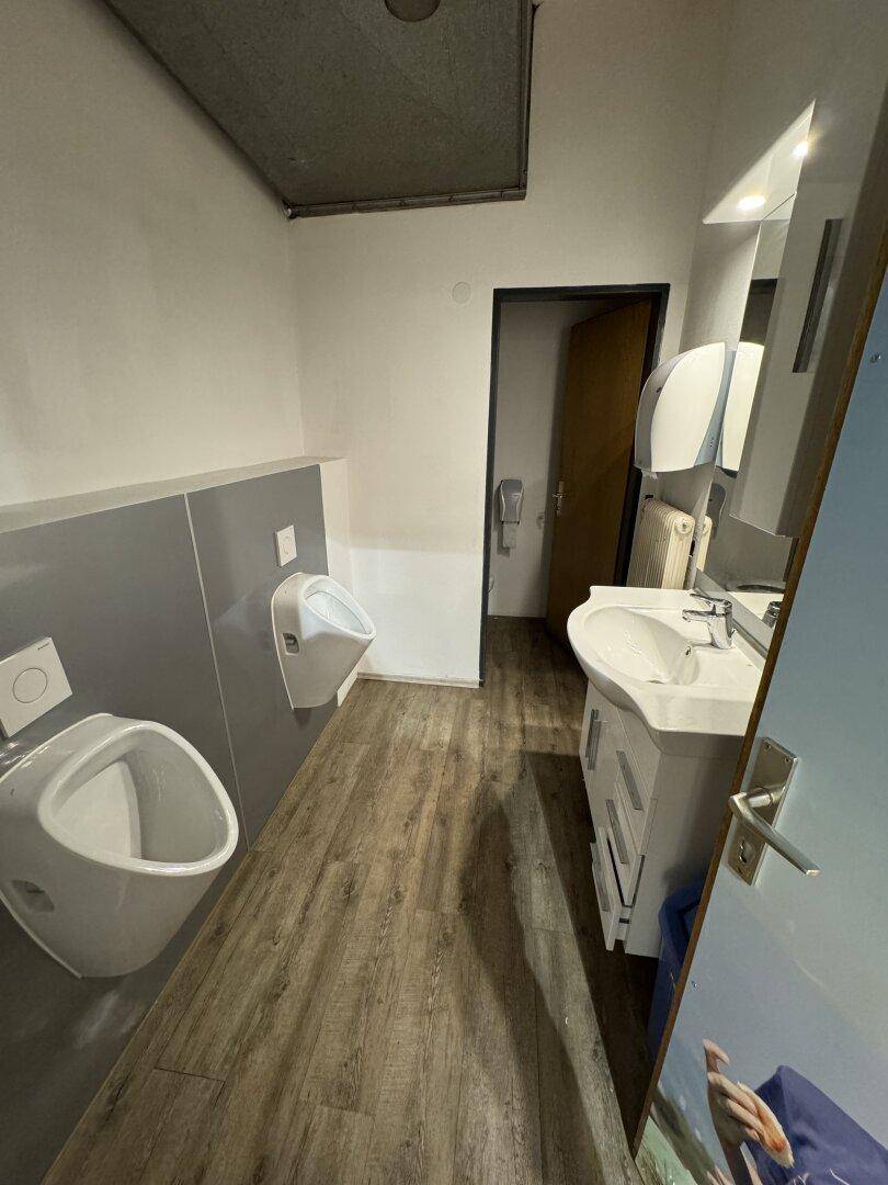 Herren-WC mit zwei Urinalen, Waschbecken und Holzboden.