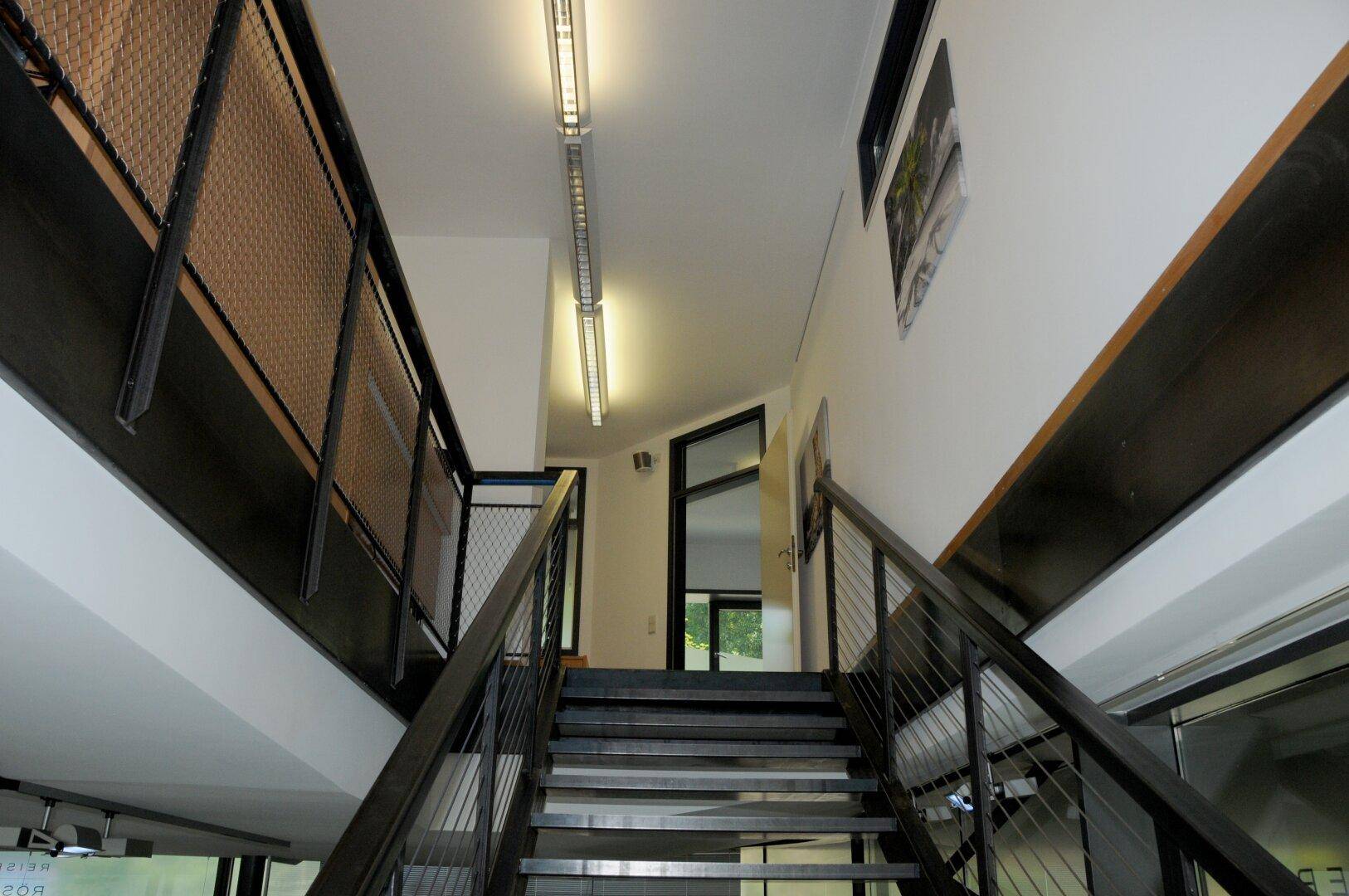 Blick nach oben auf eine moderne Metalltreppe mit schlichtem Geländer und hellen Wänden.