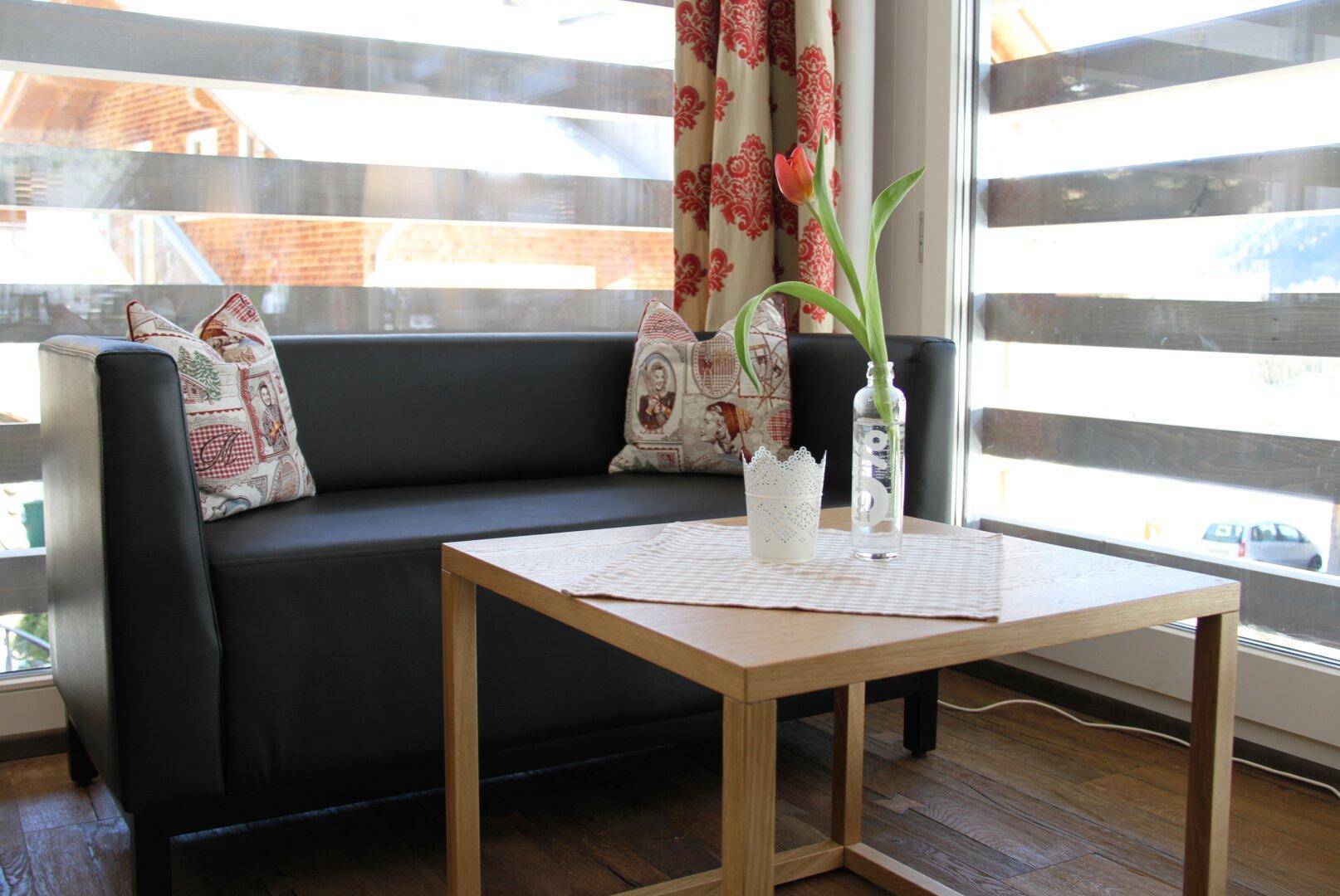 Gemütlicher kleiner Wohnbereich mit schwarzem Sofa, Holztisch und floralen Vorhängen am großen Fenster.