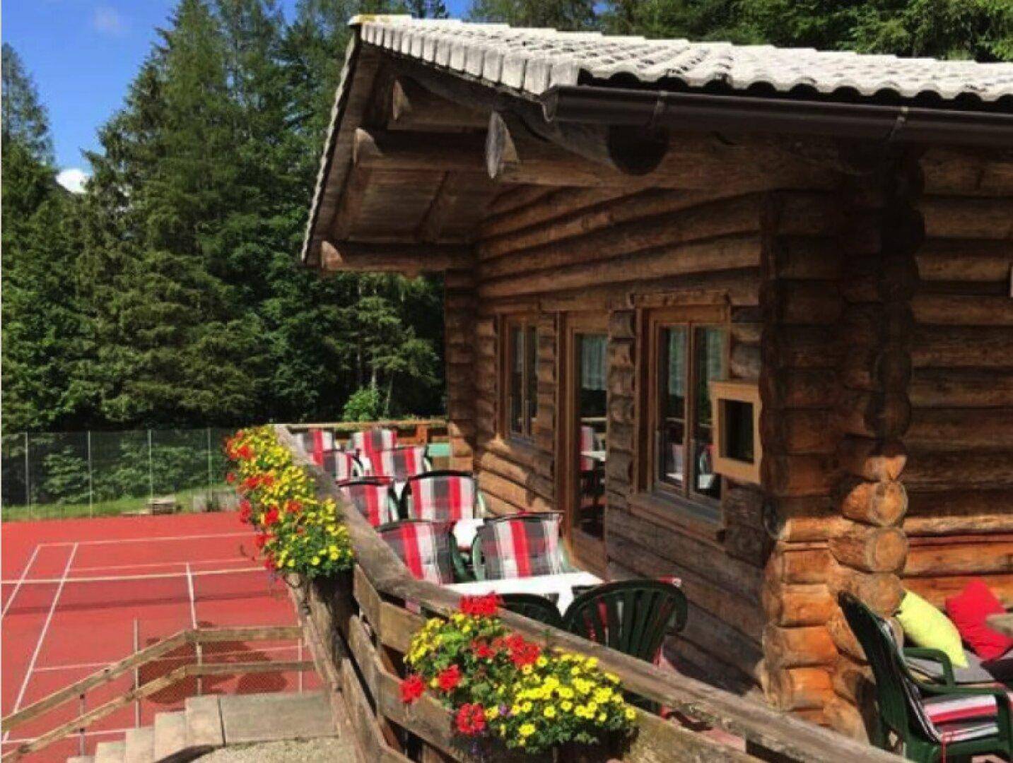 Rustikale Blockhütte mit Terrasse und Blick auf einen Tennisplatz, umgeben von Bäumen.