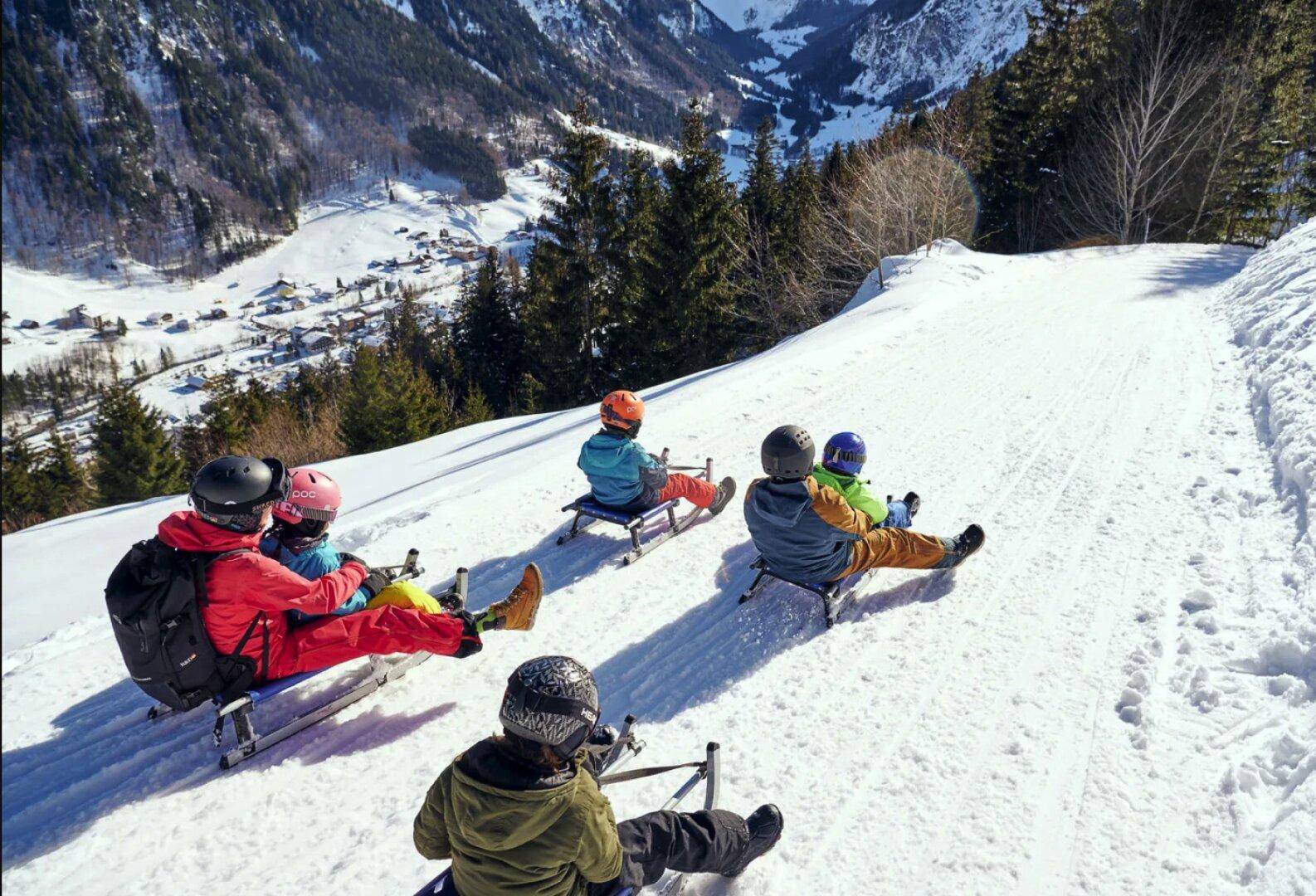 Eine Gruppe von Menschen fährt auf Schlitten einen verschneiten Hang hinunter mit Bergpanorama.