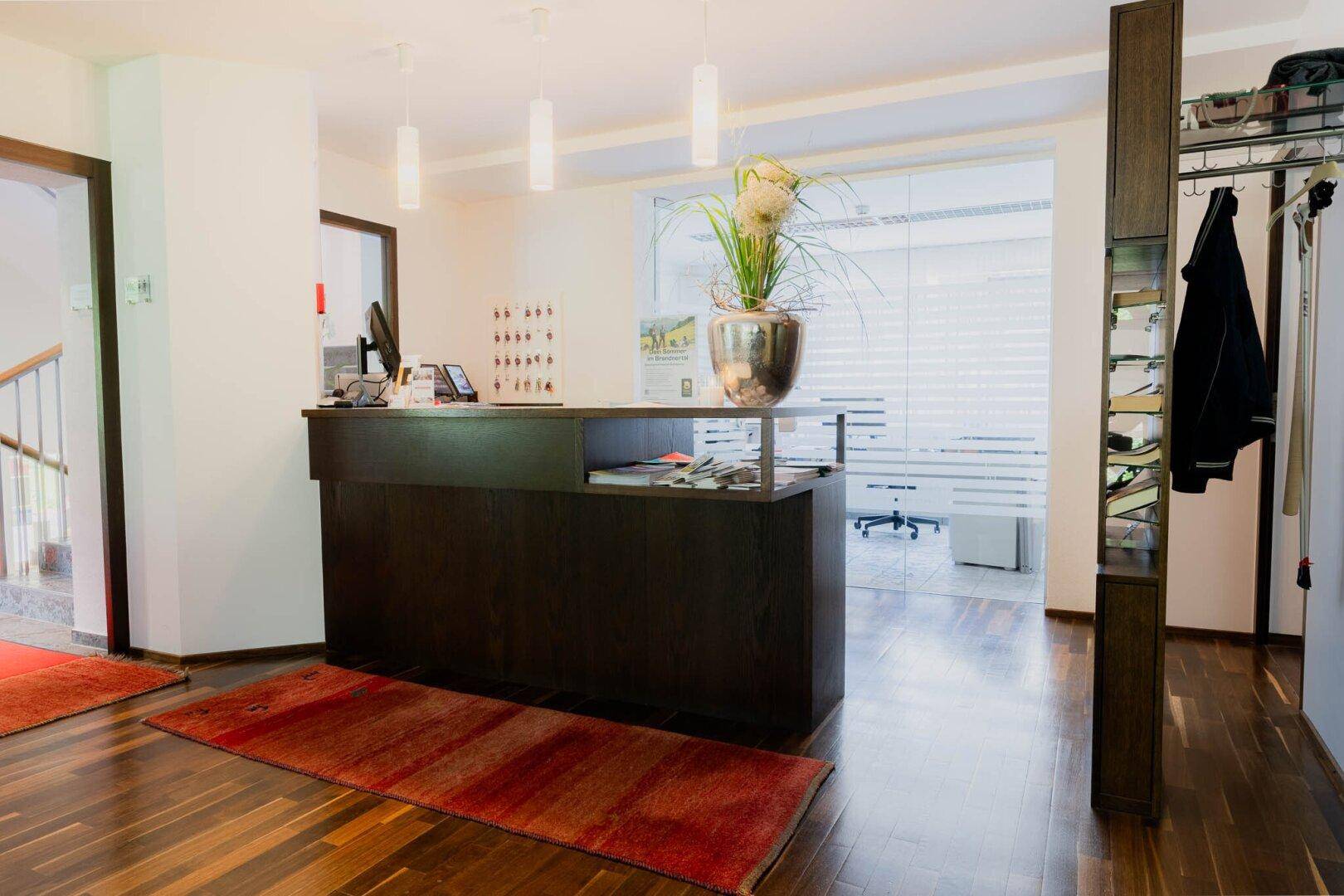 Freundlicher Empfangsbereich mit dunklem Holzschalter, Holzboden und Garderobe.