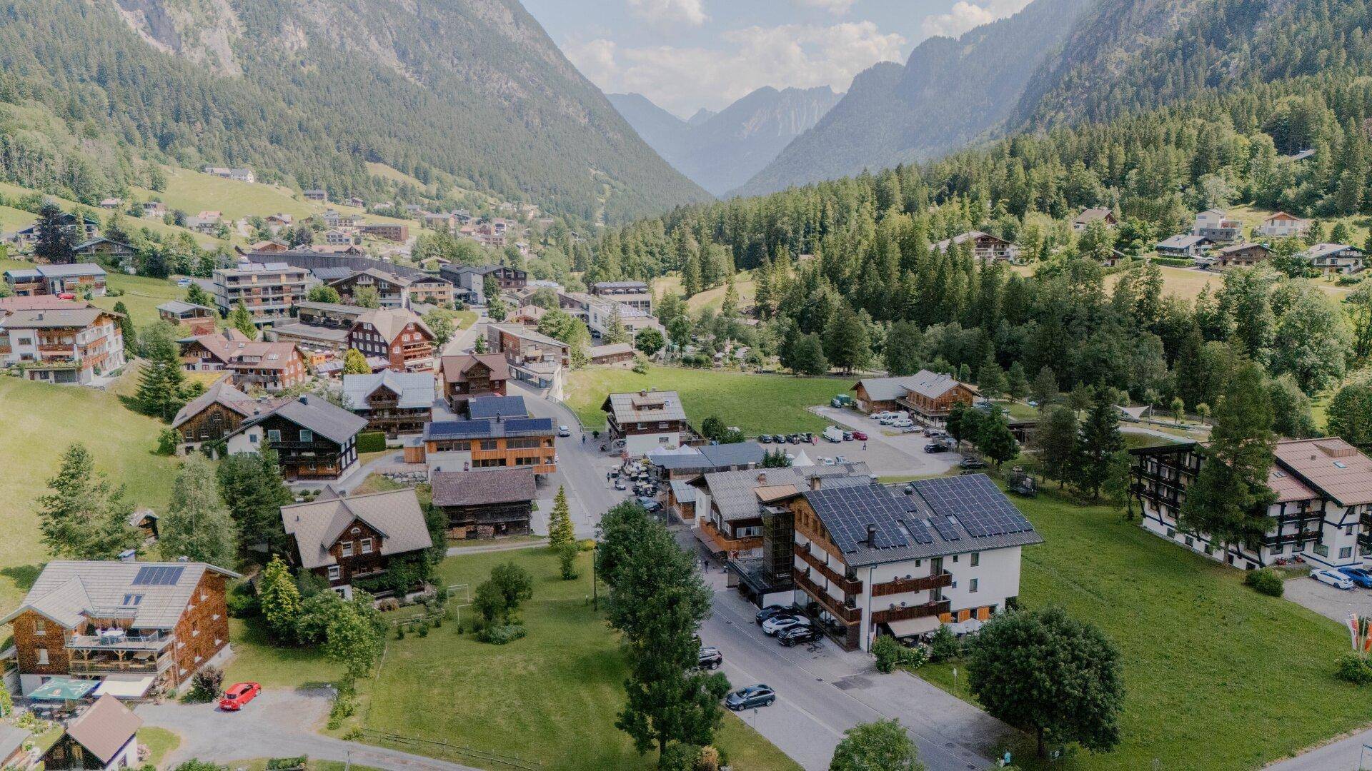 Malerisches Dorf in einem grünen Tal, umgeben von bewaldeten Bergen unter blauem Himmel.