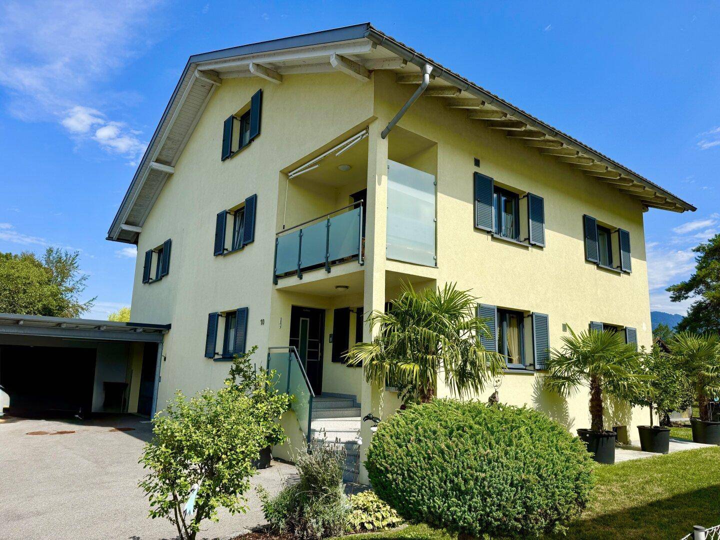Vorderansicht eines gelben Hauses mit Balkon, blauen Fensterläden und gepflegter Gartenanlage.