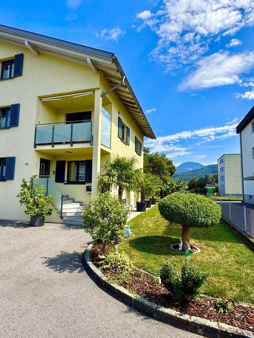 Gelbes Haus mit Balkon, gepflegtem Vorgarten und Bergblick unter strahlend blauem Himmel.