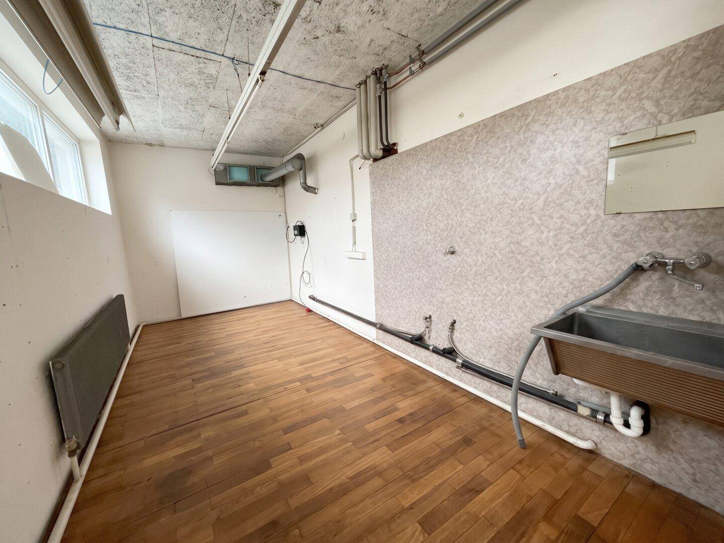 Kleiner Hauswirtschaftsraum mit Holzdielenboden, Waschbecken, sichtbaren Rohren und einem Fenster.