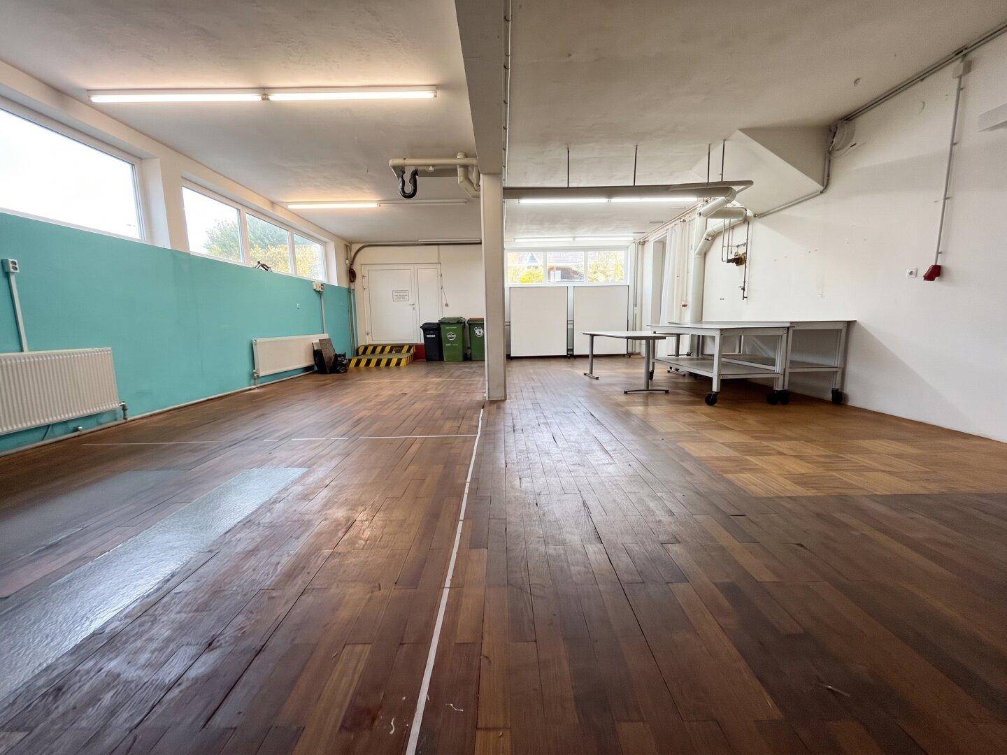 Großer Gewerberaum mit Holzdielenboden, farbiger Wand und Fenstern, ausgestattet mit Tischen und Regalen.