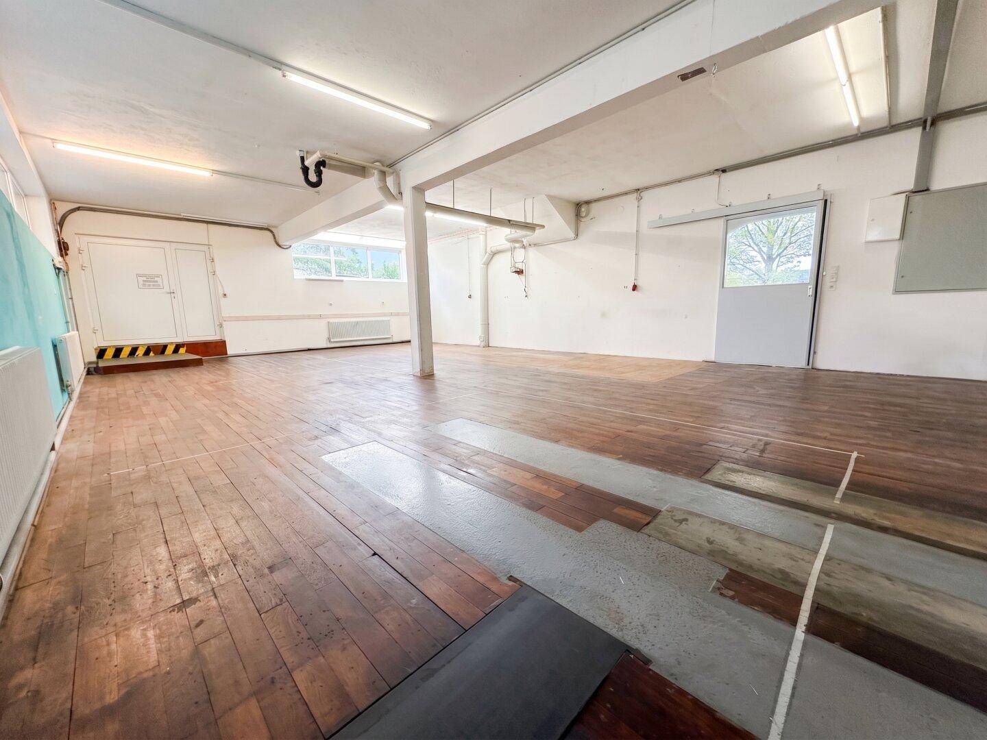 Großer, leerer Gewerberaum mit Holzdielenboden, sichtbaren Rohren und Leuchtstoffröhrenbeleuchtung.