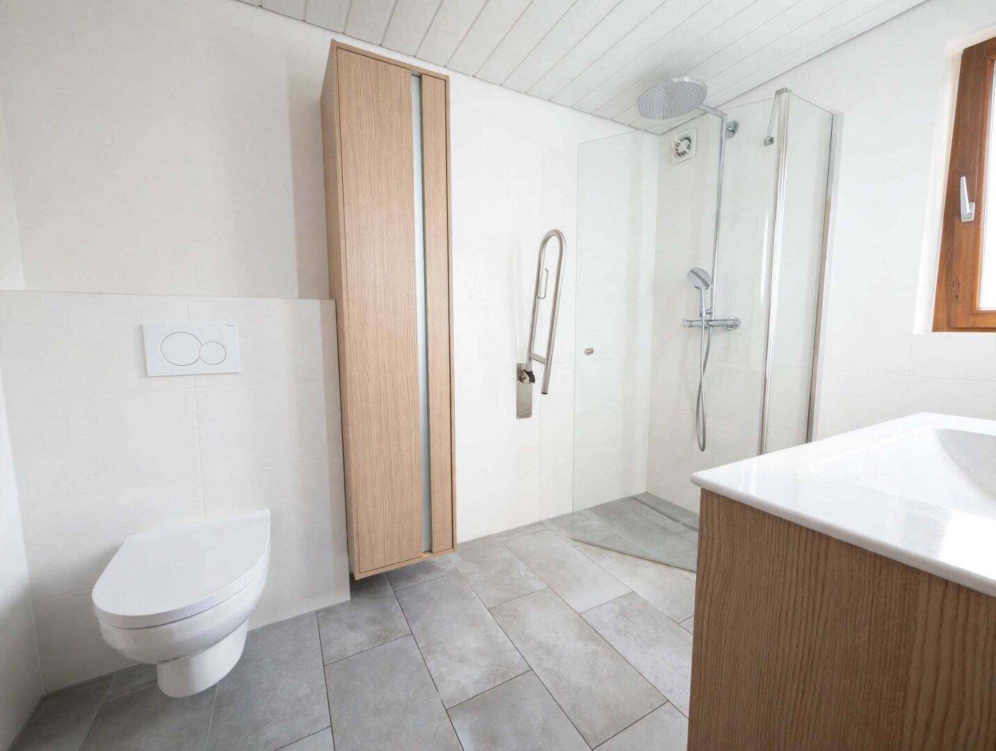 Helles Badezimmer mit wandhängendem WC, ebenerdiger Dusche und Haltegriffen, auf hellgrauen Fliesen.