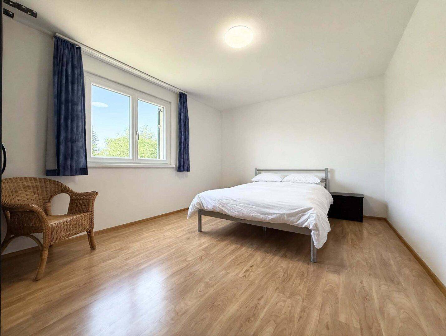 Helles Schlafzimmer mit einfachem Bett, Korbstuhl und hellem Holzboden, mit Blick ins Grüne.