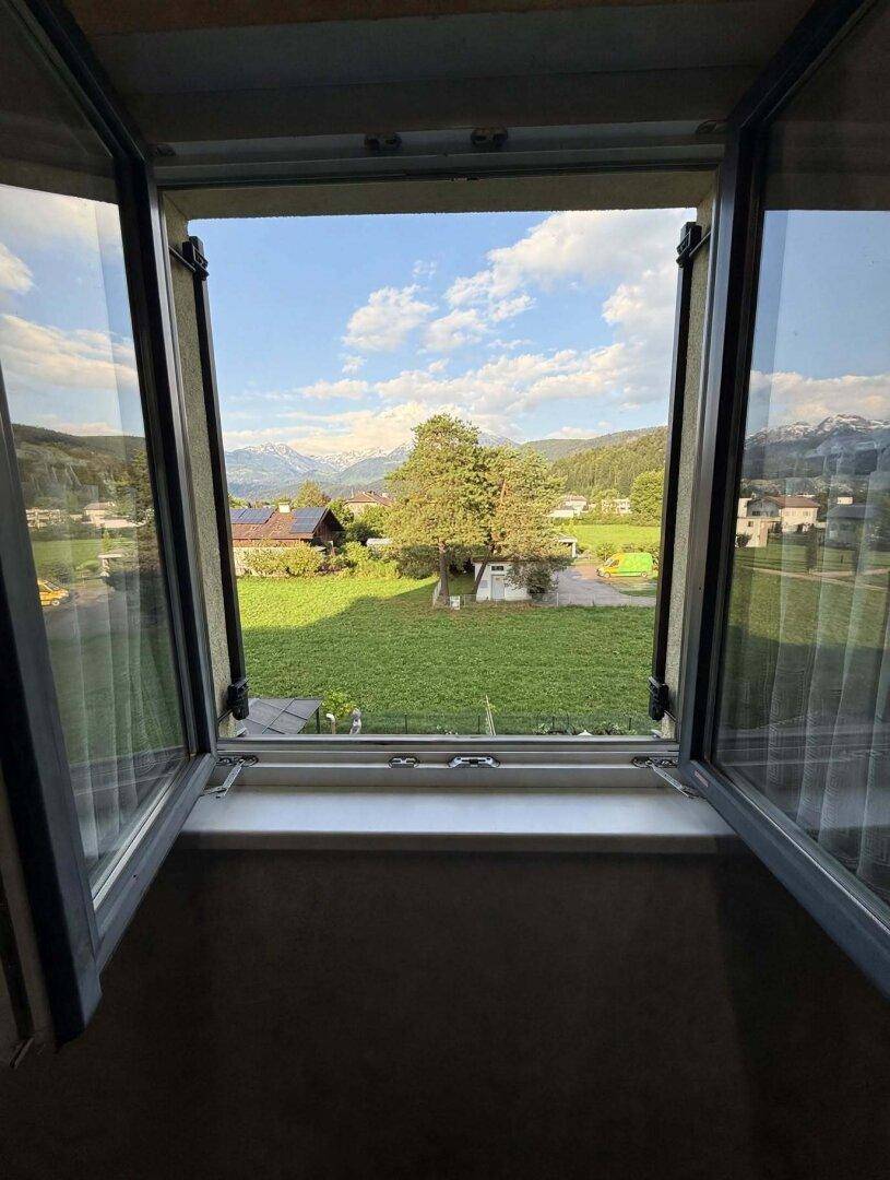 Fensterblick auf eine grüne Landschaft mit Häusern und Bergen in der Ferne, bietet eine weite Aussicht.