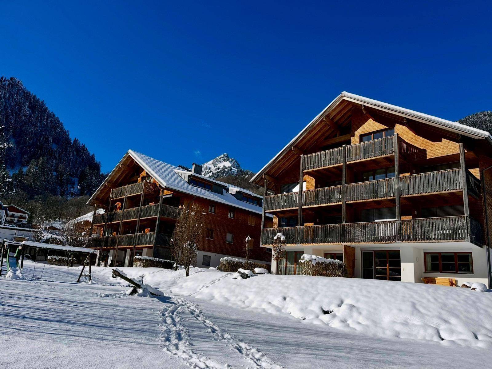 Zwei moderne Chalet-Gebäude mit Balkonen und schneebedeckter Umgebung an einem sonnigen Wintertag.