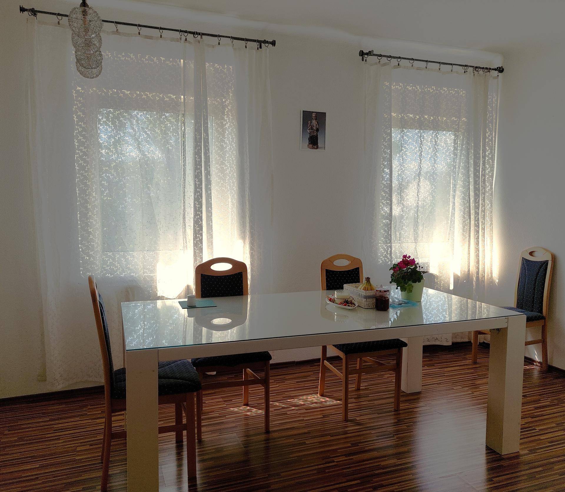 Heller Essbereich mit großem Tisch und Stühlen, beleuchtet durch zwei Fenster mit Vorhängen.