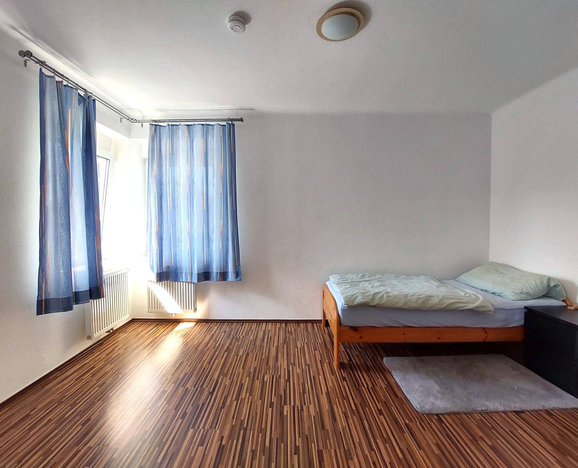 Gemütliches Schlafzimmer mit Einzelbett, Laminatboden und einem Fenster mit blauen Vorhängen.