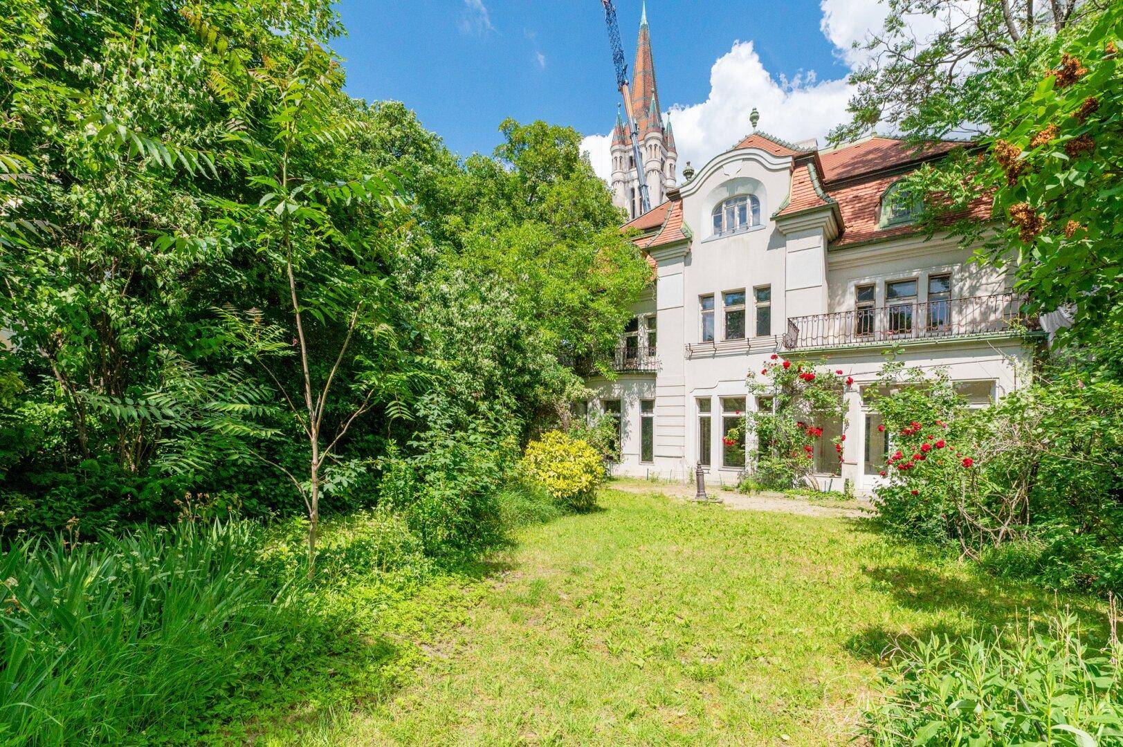 Historisches Gebäude mit verwildertem Garten und einem Kirchturm, umgeben von üppigem Grün.