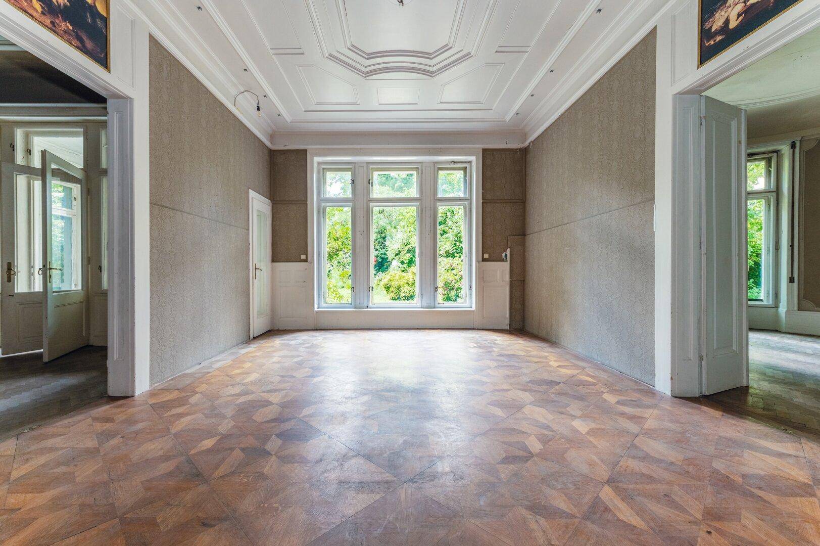 Geräumiges Zimmer mit Fischgrätparkett, hohen Decken und Stuckverzierungen, hell durch große Fenster.