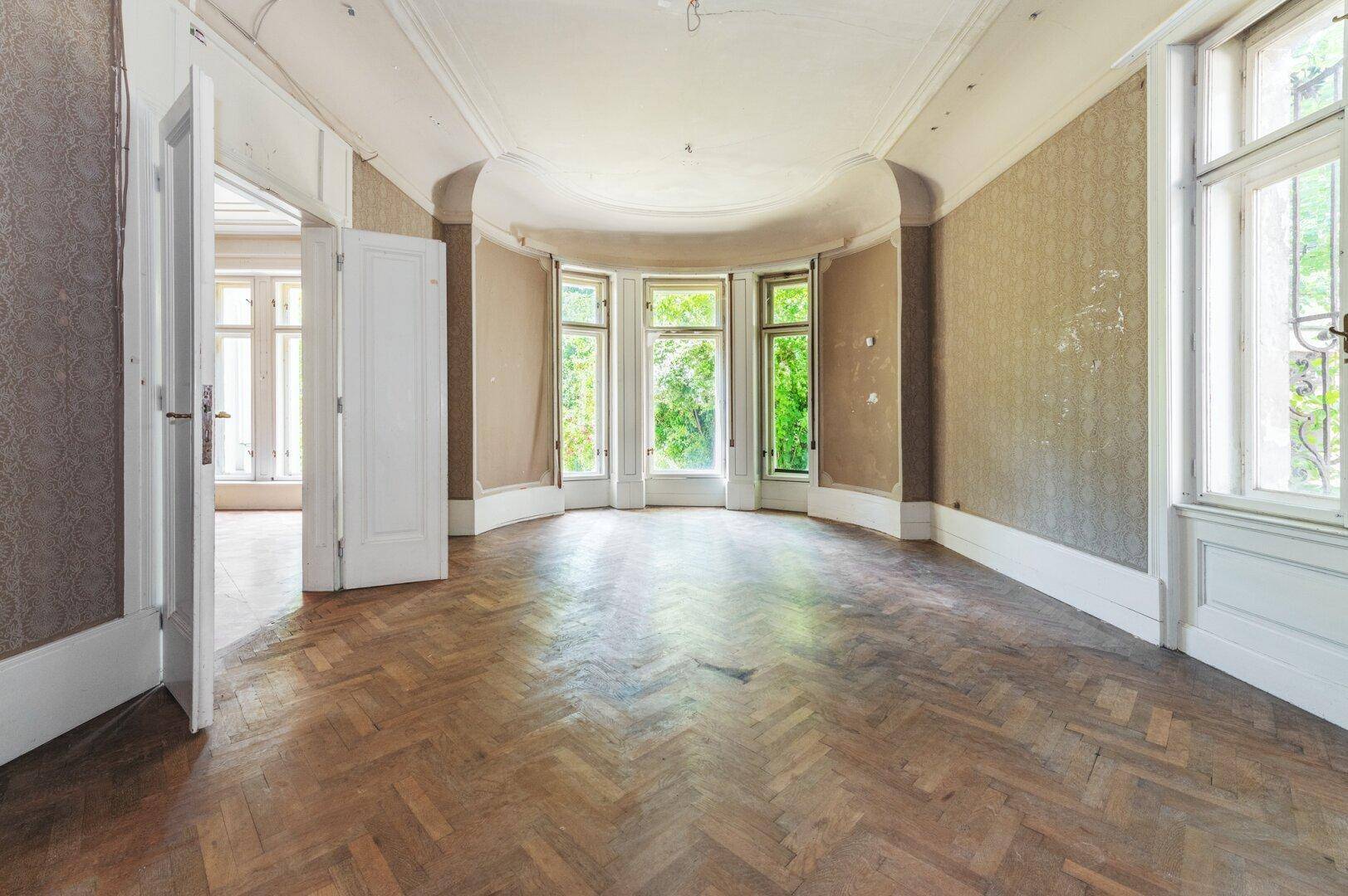 Helles Zimmer mit Erkerfenstern, Fischgrätparkett und Blick ins Grüne, ideal für einen Salon.