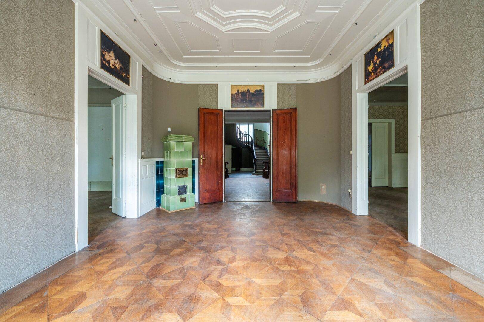 Historisches Zimmer mit Fischgrätparkett, grünem Kachelofen und hohen Decken, bereit zur Renovierung.