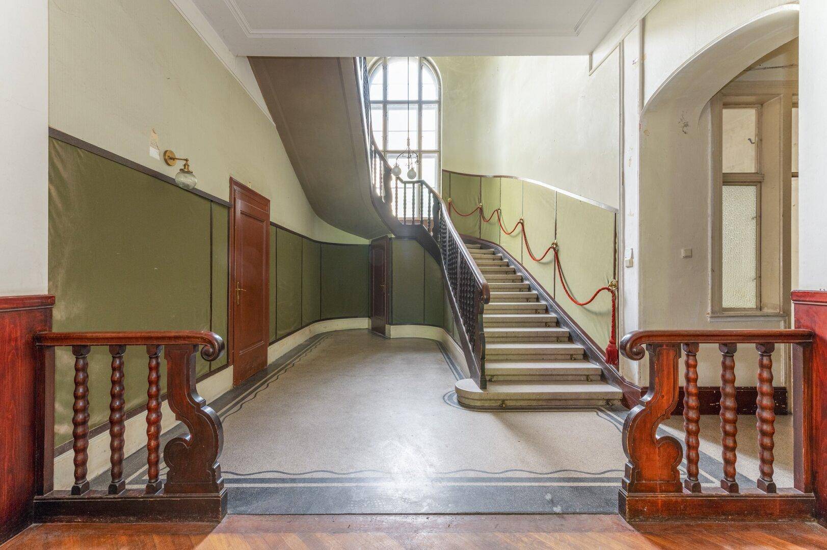 Eindrucksvolles Treppenhaus mit geschwungener Holztreppe und hohen Wänden in einem historischen Gebäude.