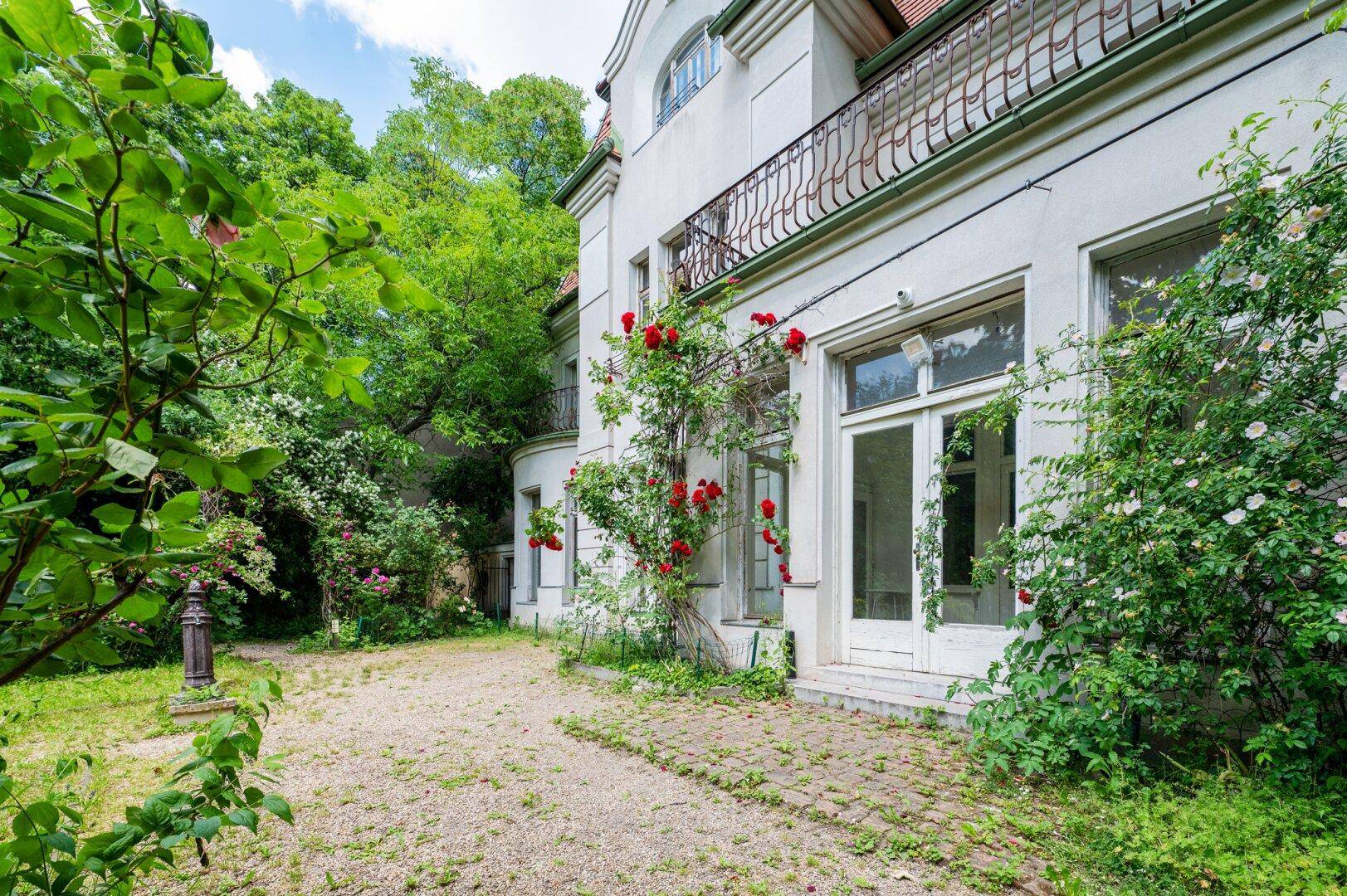 Rückseite des Gebäudes mit verwildertem Garten und blühenden roten Rosensträuchern.