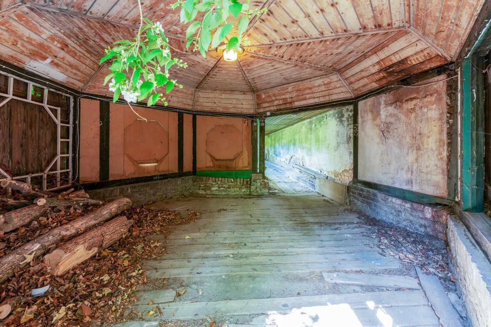 Verfallenes Gartenhaus oder Pavillon mit Holzdecke und gelagertem Holz, umgeben von Laub.