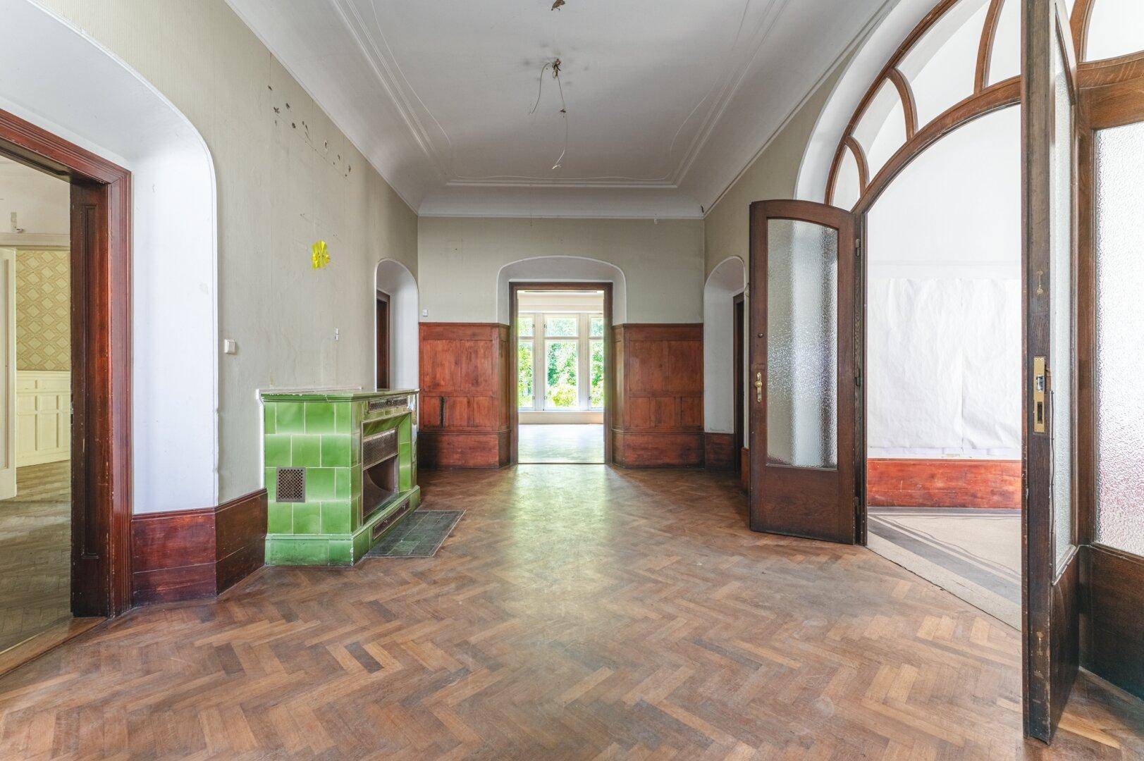 Geräumiges Zimmer mit Fischgrätparkett, grünem Kachelofen und Holztäfelung, mit Blick in den Garten.
