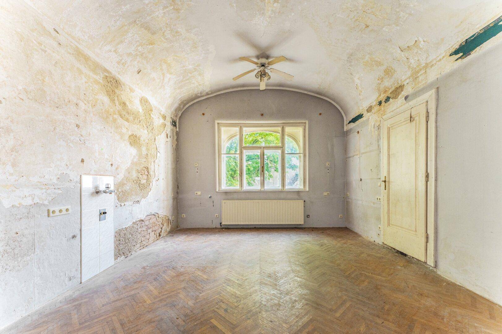 Renovierungsbedürftiger Raum mit gewölbter Decke, abgenutztem Parkettboden und großem Fenster.