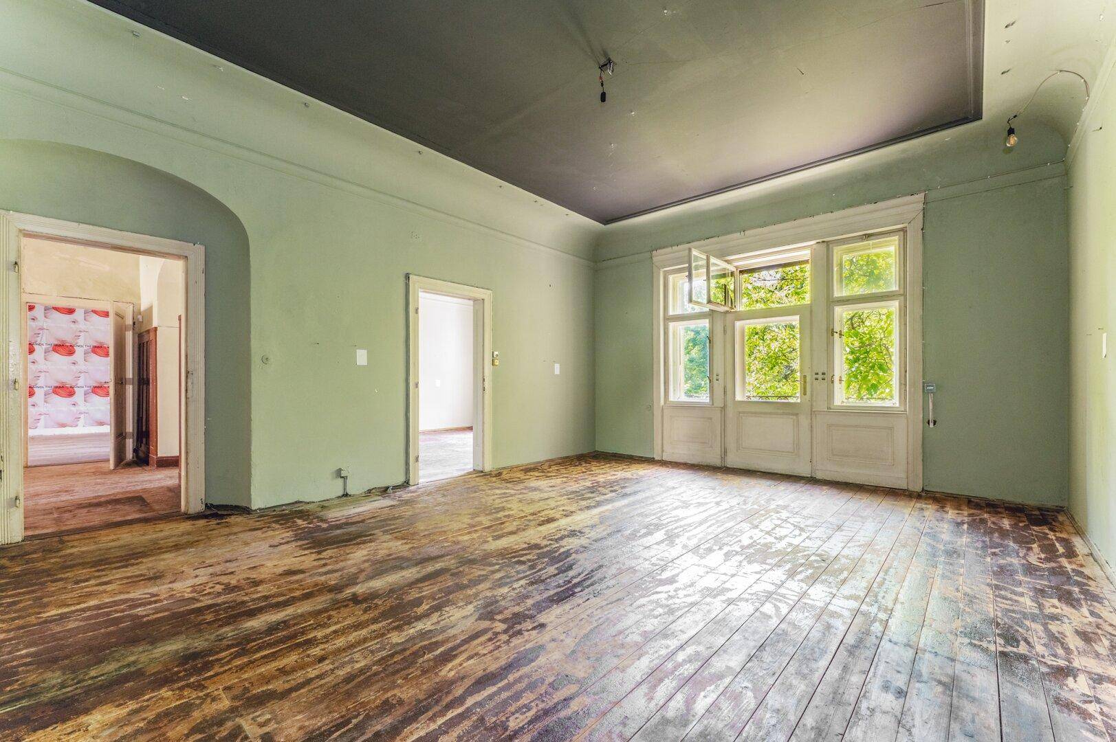Geräumiges Zimmer mit abgenutztem Holzboden, hellgrünen Wänden und Blick ins Grüne durch große Fenster.