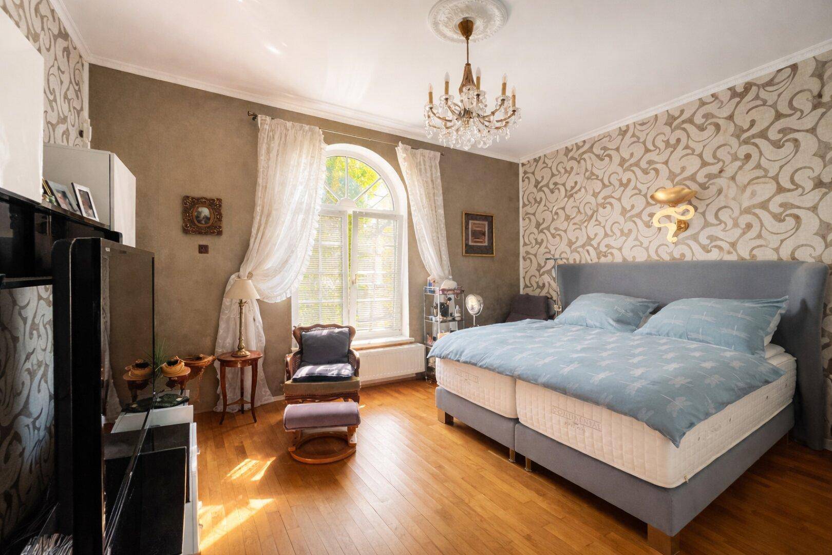 Helles Schlafzimmer mit Parkettboden, gemusterter Tapete und einem großen Bogenfenster.