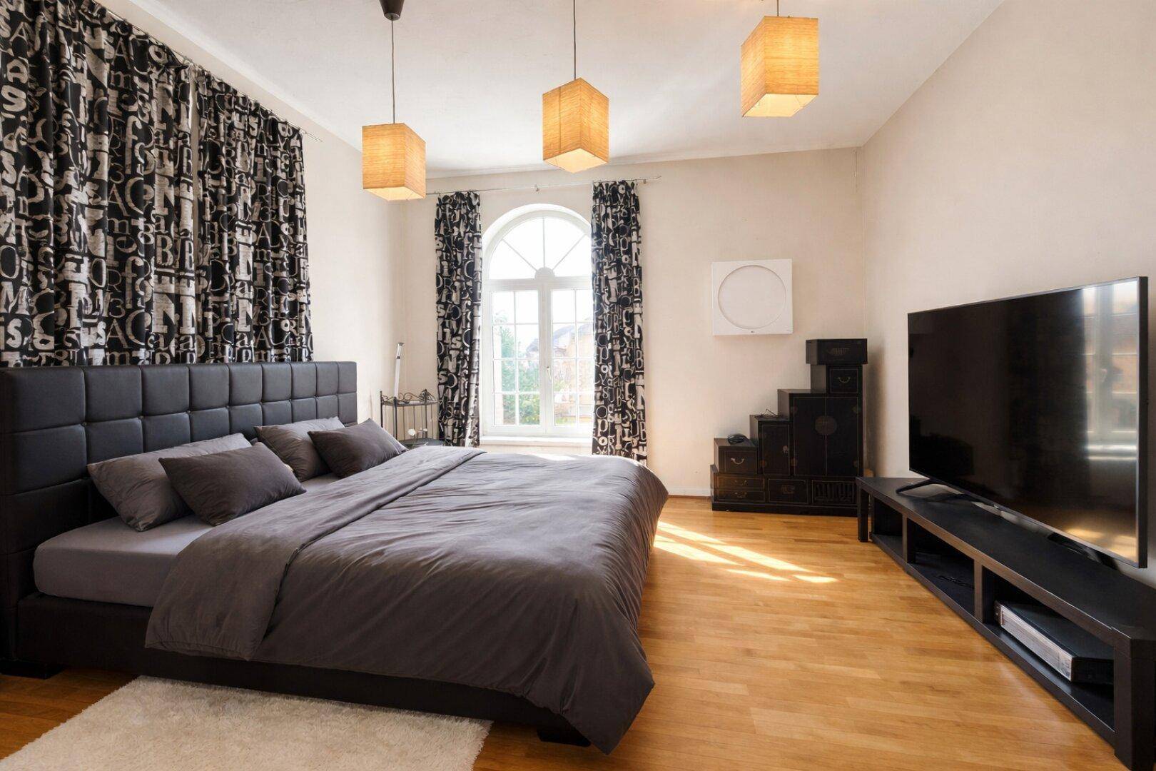 Modern eingerichtetes Schlafzimmer mit Holzboden, großem Bett und dunklen Vorhängen.