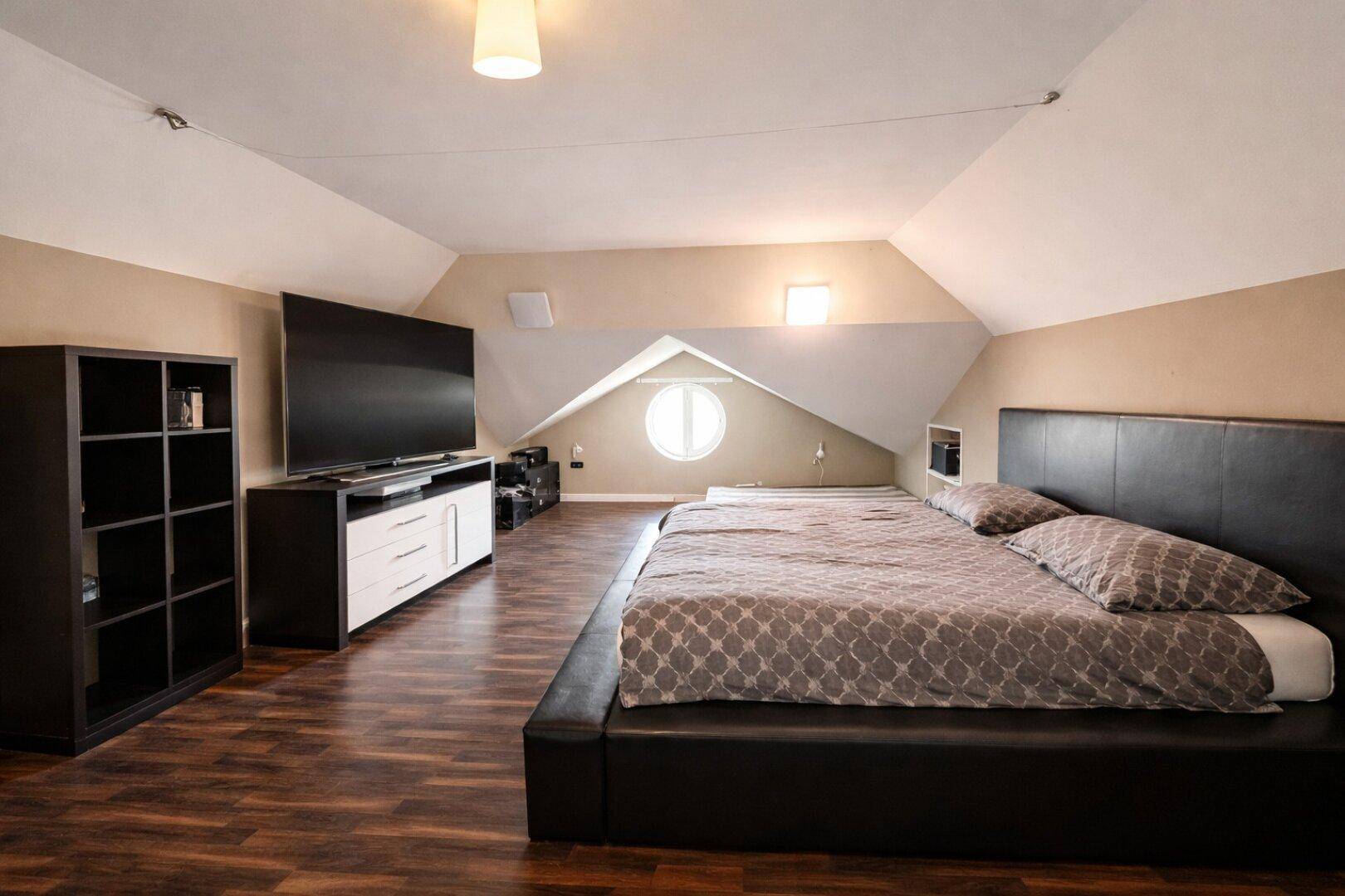 Geräumiges Schlafzimmer im Dachgeschoss mit Holzboden, großem Bett und runden Fenstern.