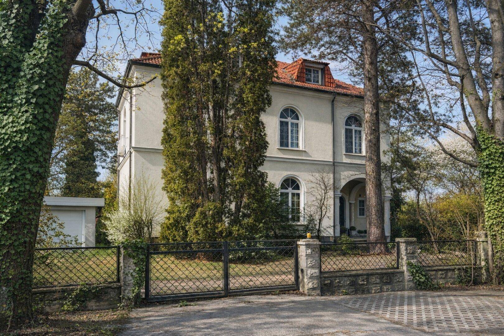 Repräsentative Villa mit klassischer Fassade, umgeben von altem Baumbestand und einem Zaun.