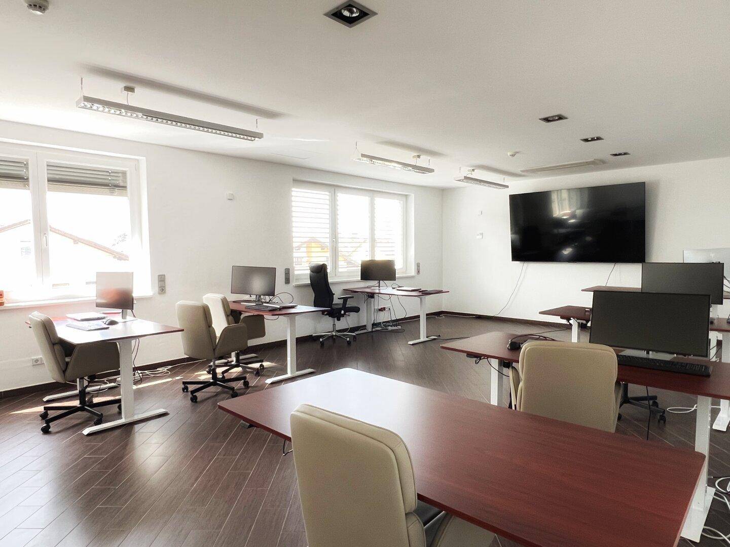 Großes Büro mit mehreren Arbeitsplätzen, Holzboden und großen Fenstern.