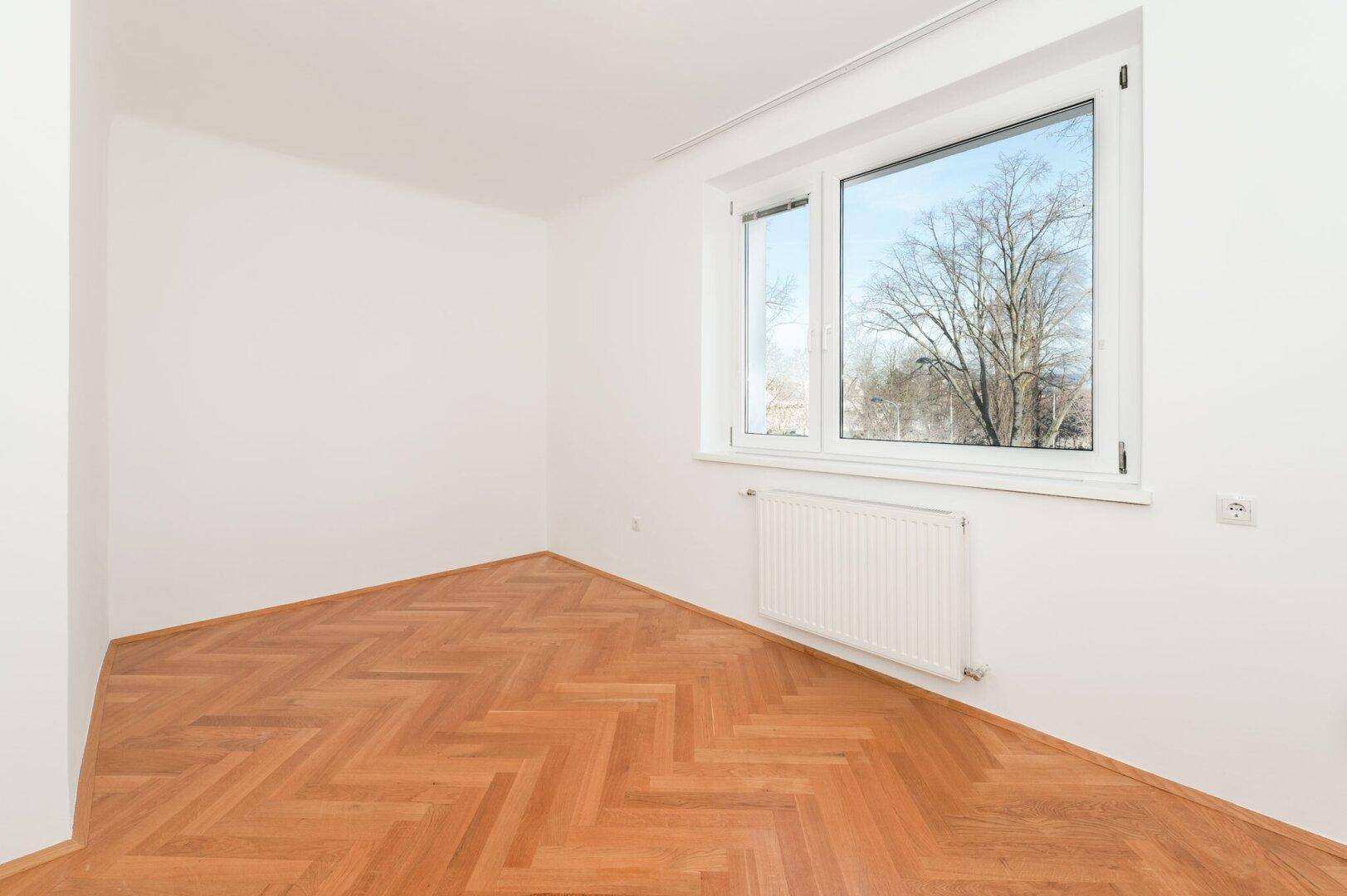 Leeres Zimmer mit hellem Fischgrätparkett und großem Fenster, das eine angenehme Helligkeit bietet.