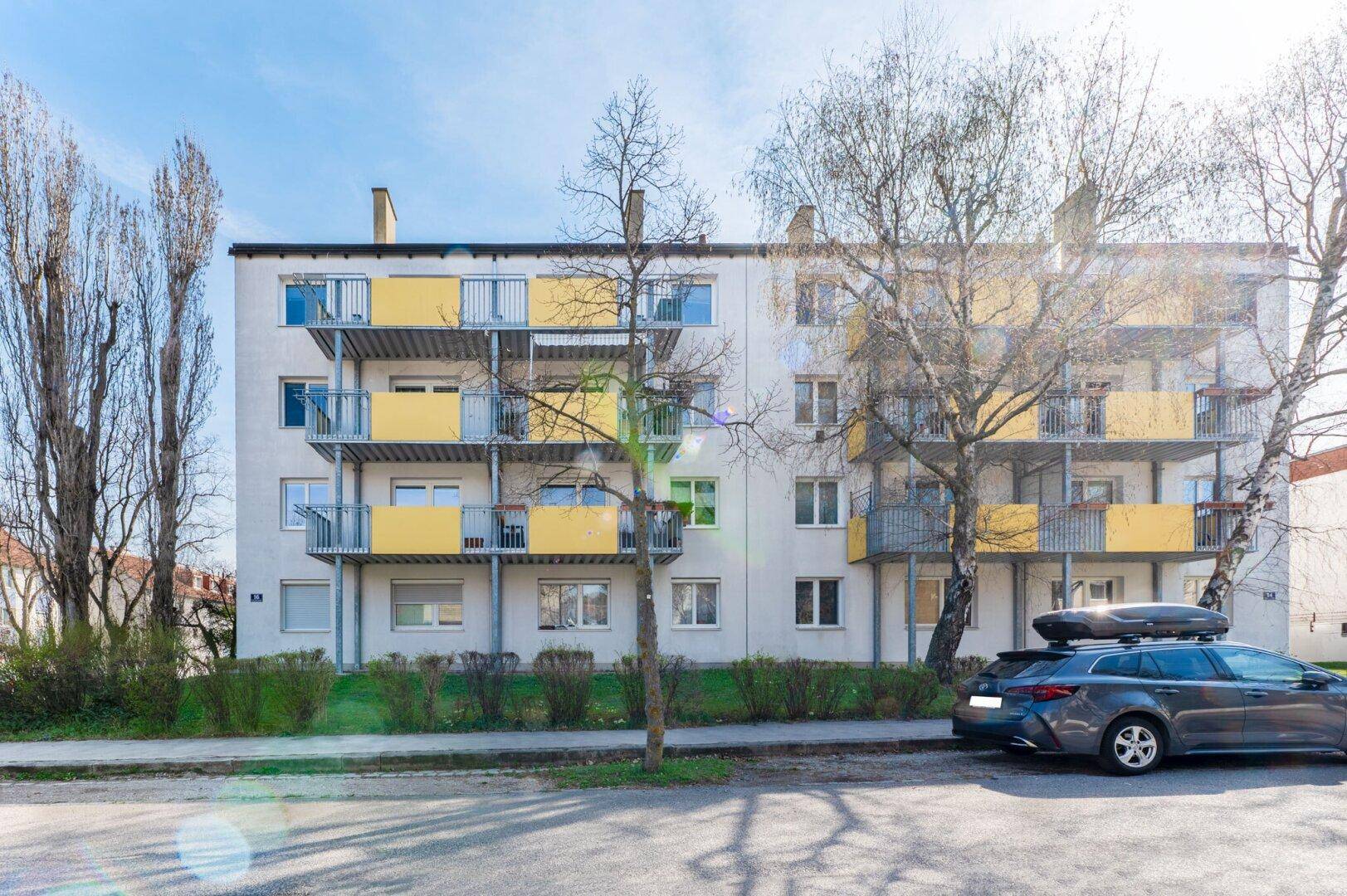 Mehrfamilienhaus mit gelben Balkonen und Bäumen an einer sonnigen Straße, gut gepflegt.