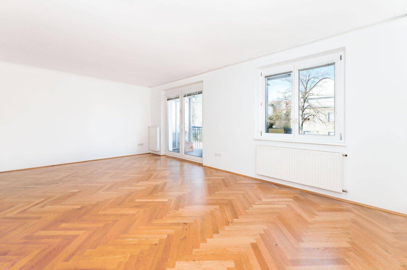Helles Wohnzimmer mit Fischgrätparkettboden und Zugang zu einem Balkon, das viel Tageslicht bietet.