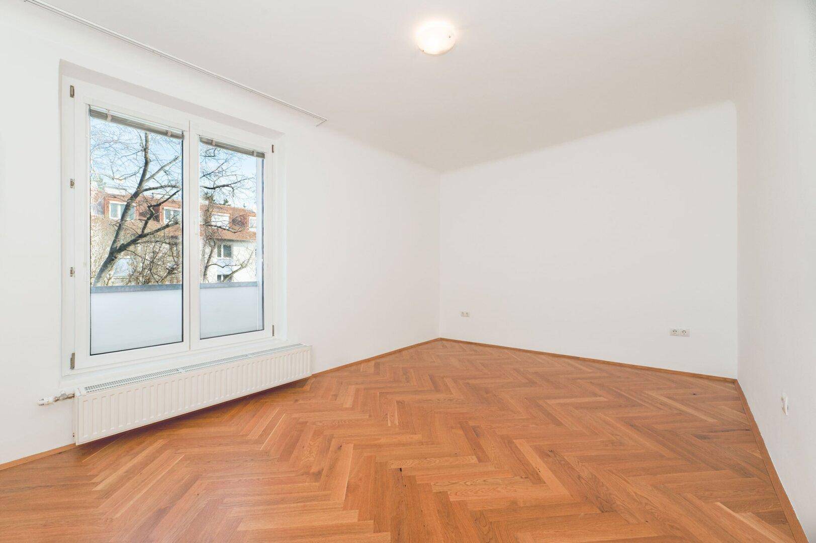 Leeres Zimmer mit hellem Fischgrätparkett und großem Fenster, das einen Blick ins Grüne ermöglicht.