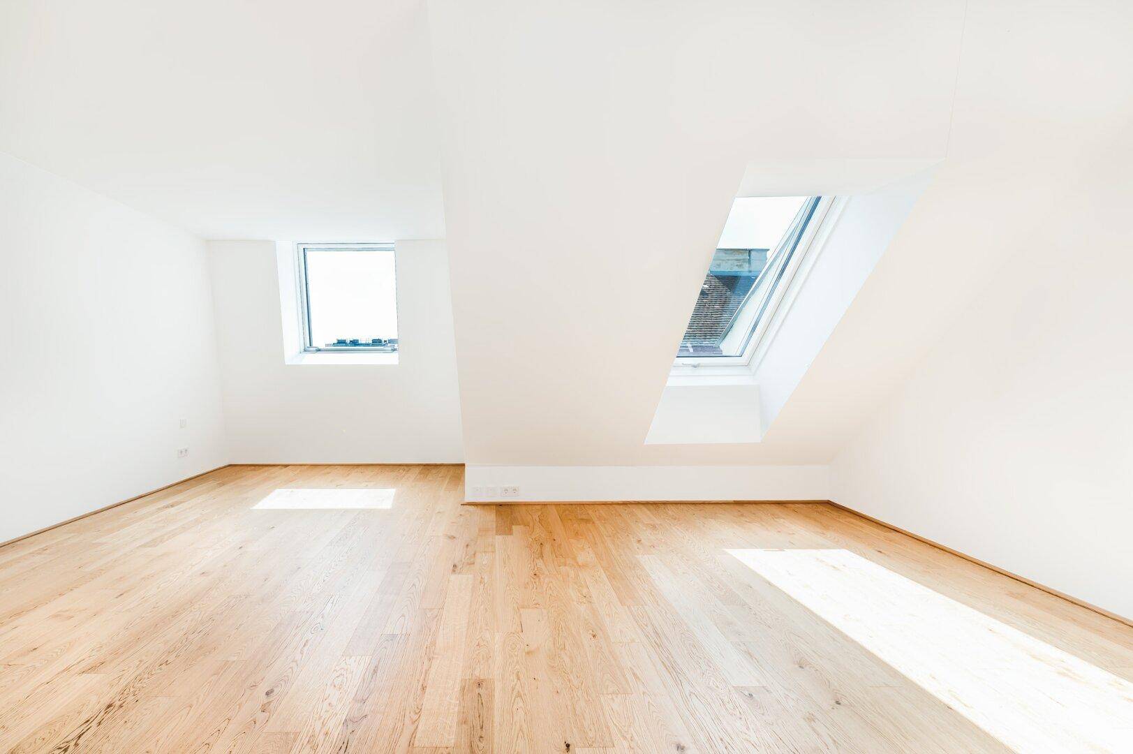 Helles Schlafzimmer mit Holzboden, weißen Wänden und zwei Fenstern, darunter ein Dachfenster.