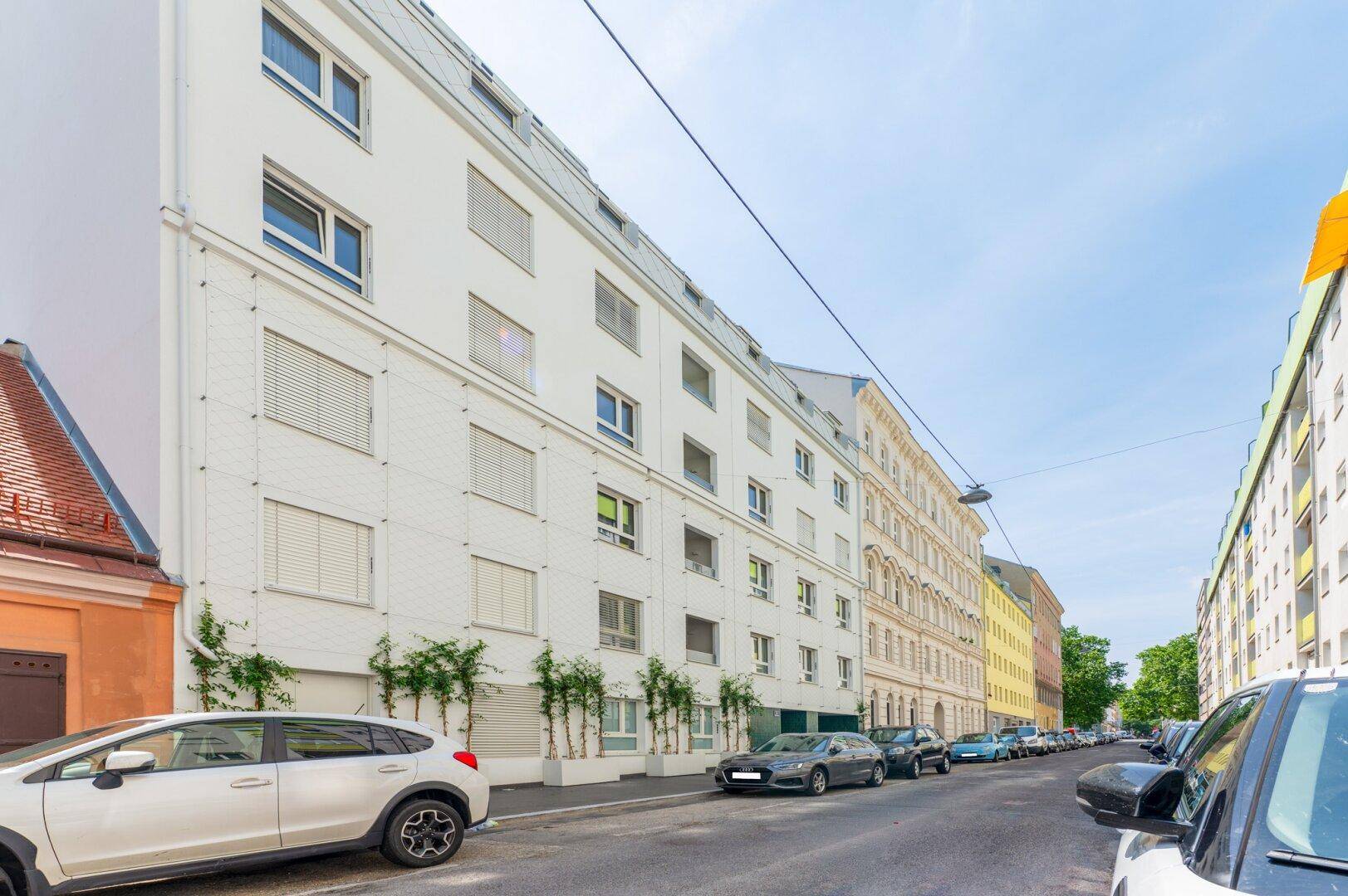 Moderne weiße Wohnhausfassade an einer belebten Straße mit parkenden Autos und klarem Himmel.