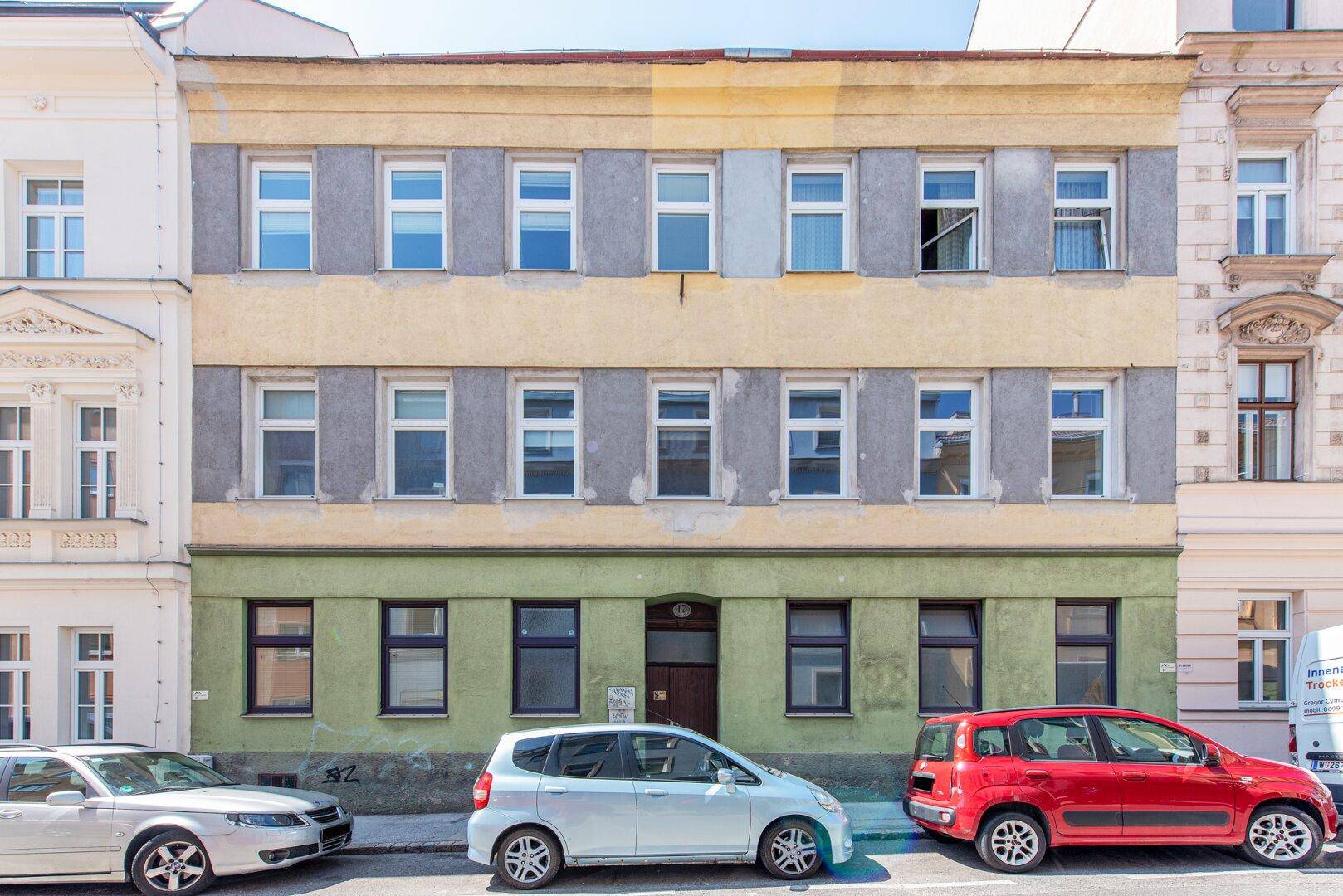 Ältere Gebäudefassade mit sichtbaren Gebrauchsspuren und parkenden Autos auf der Straße.
