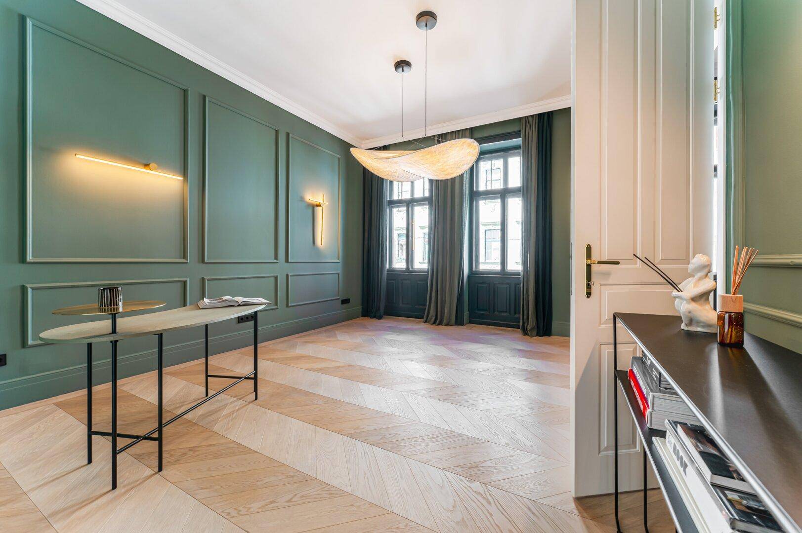 Geräumiges Wohnzimmer mit grünen Wandpaneelen, hohen Fenstern und elegantem Fischgrätparkett.