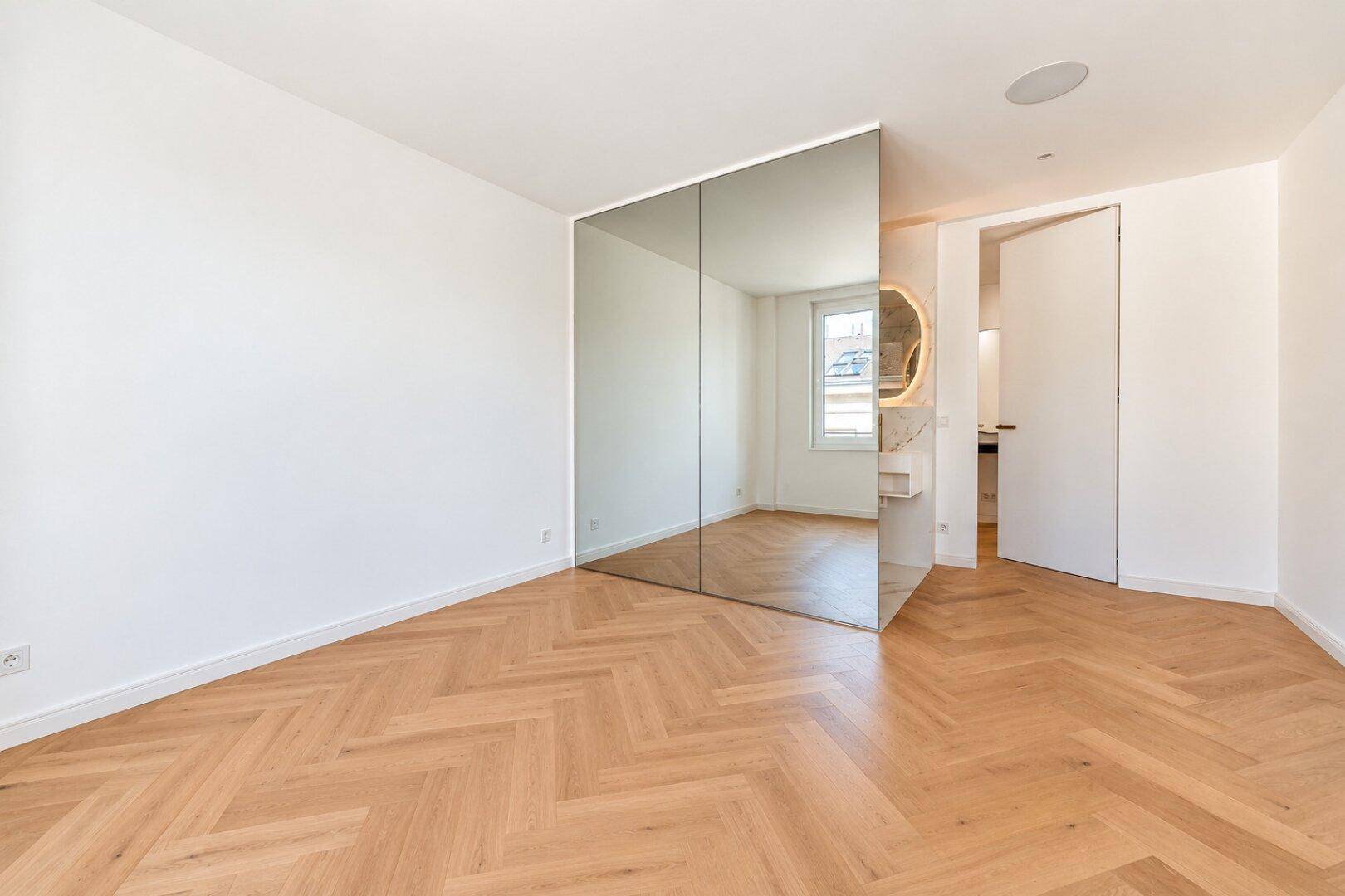 Helles Schlafzimmer mit Fischgrätparkett, großer Spiegelwand und Zugang zum Badezimmer.