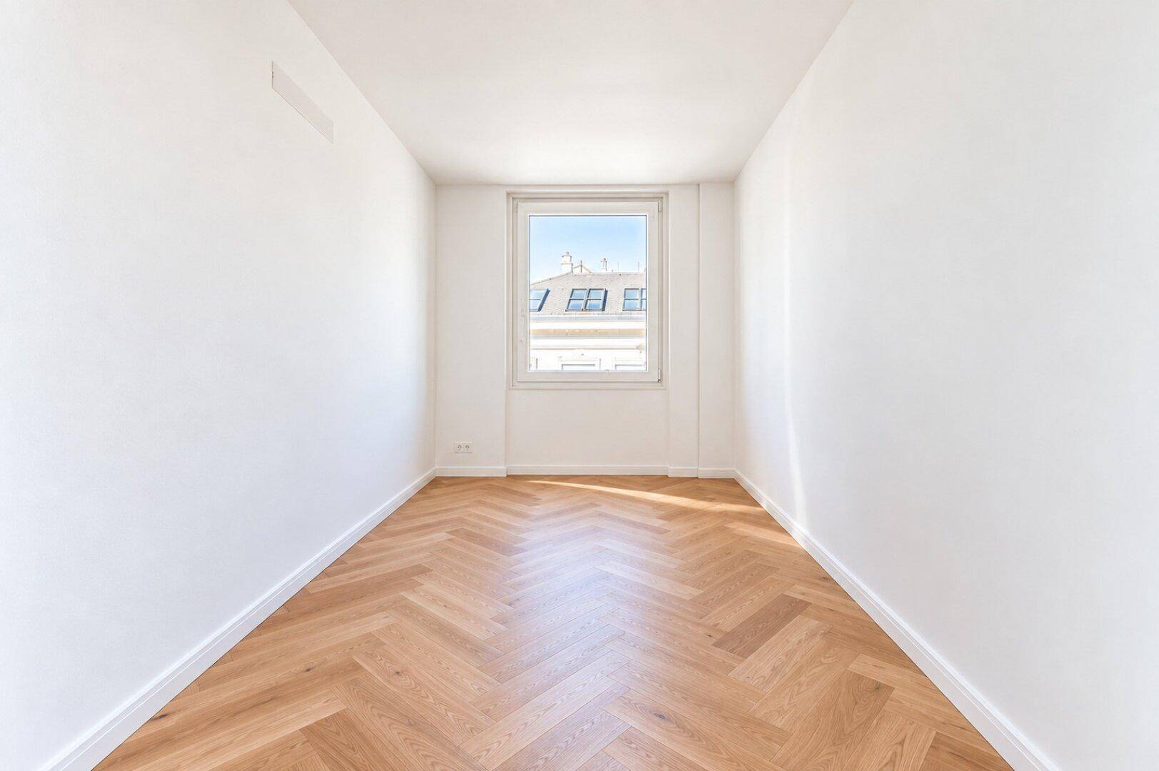 Helles, leeres Zimmer mit Fischgrätparkett und einem Fenster mit Blick nach draußen.