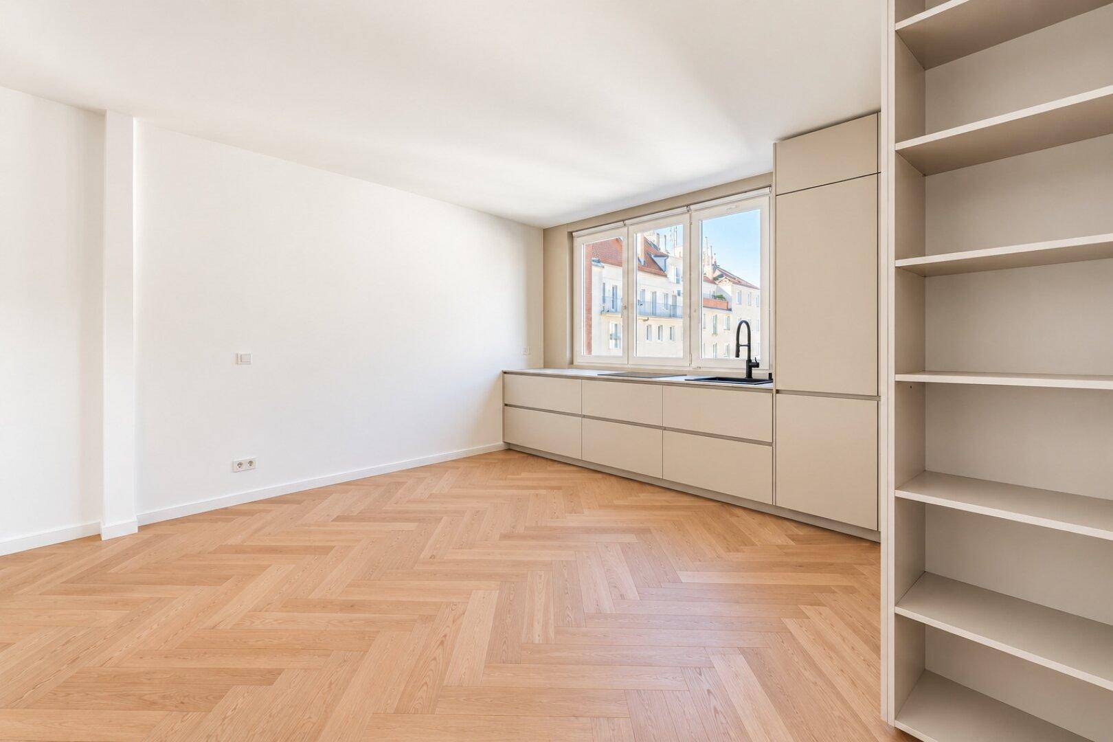 Helle Küche mit integrierten Schränken, Fenster und Fischgrätparkett in einem geräumigen, leeren Raum.