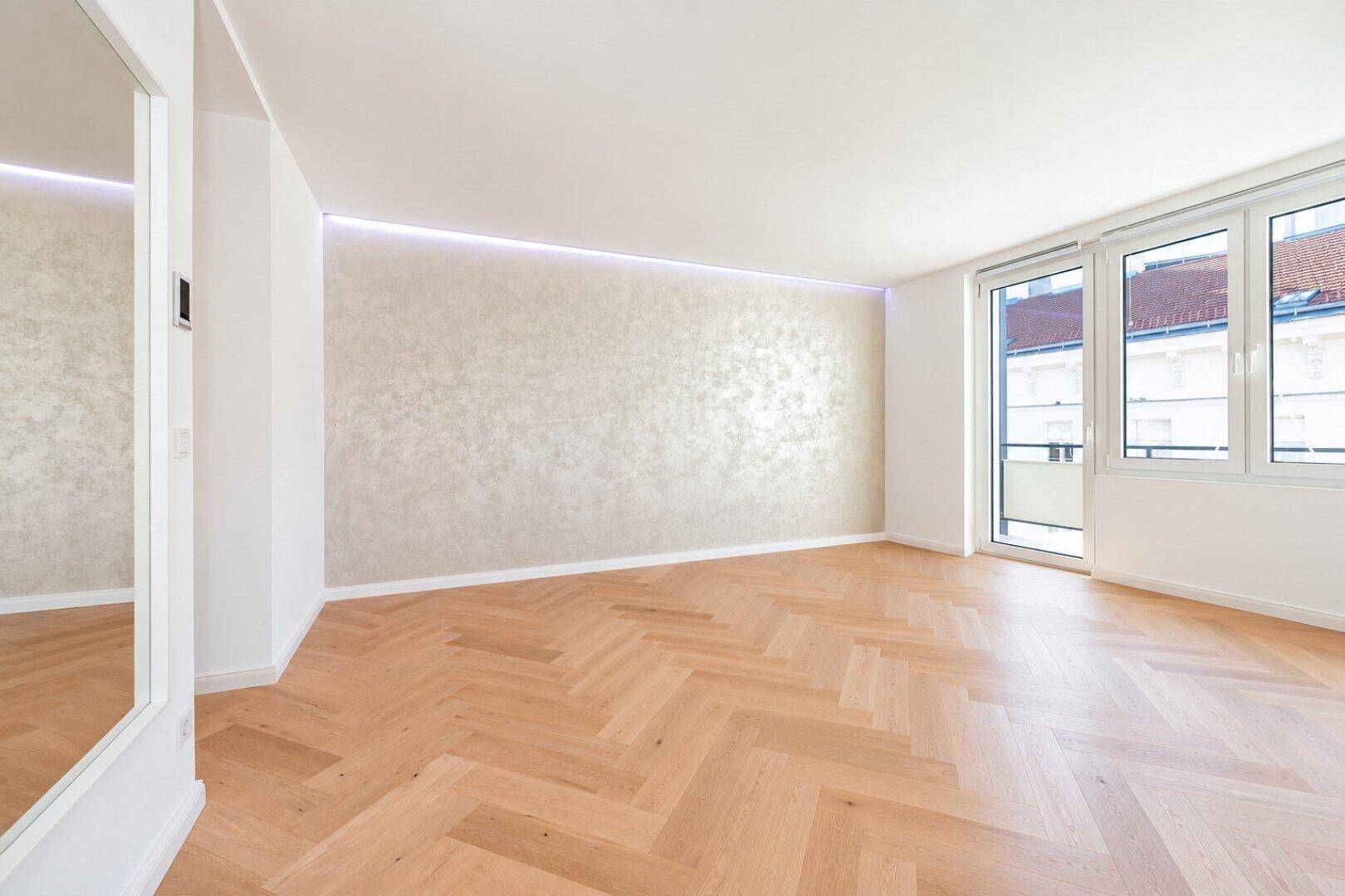 Geräumiges Wohnzimmer mit Fischgrätparkett, heller Akzentwand und Zugang zu einem Balkon.