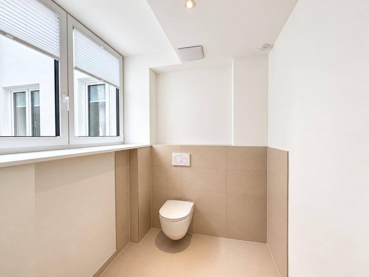 Separate Toilette mit wandhängendem WC, Fenster und hellen Fliesen.
