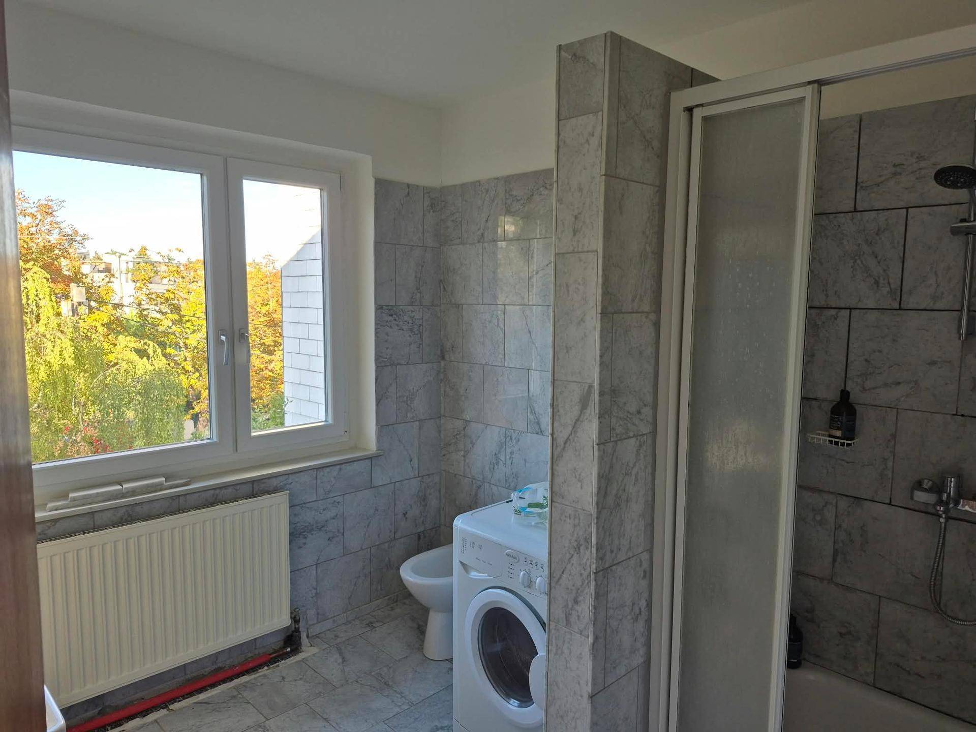 Badezimmer mit Duschkabine, Waschmaschine, gefliesten Wänden und einem Fenster für natürliche Belüftung.
