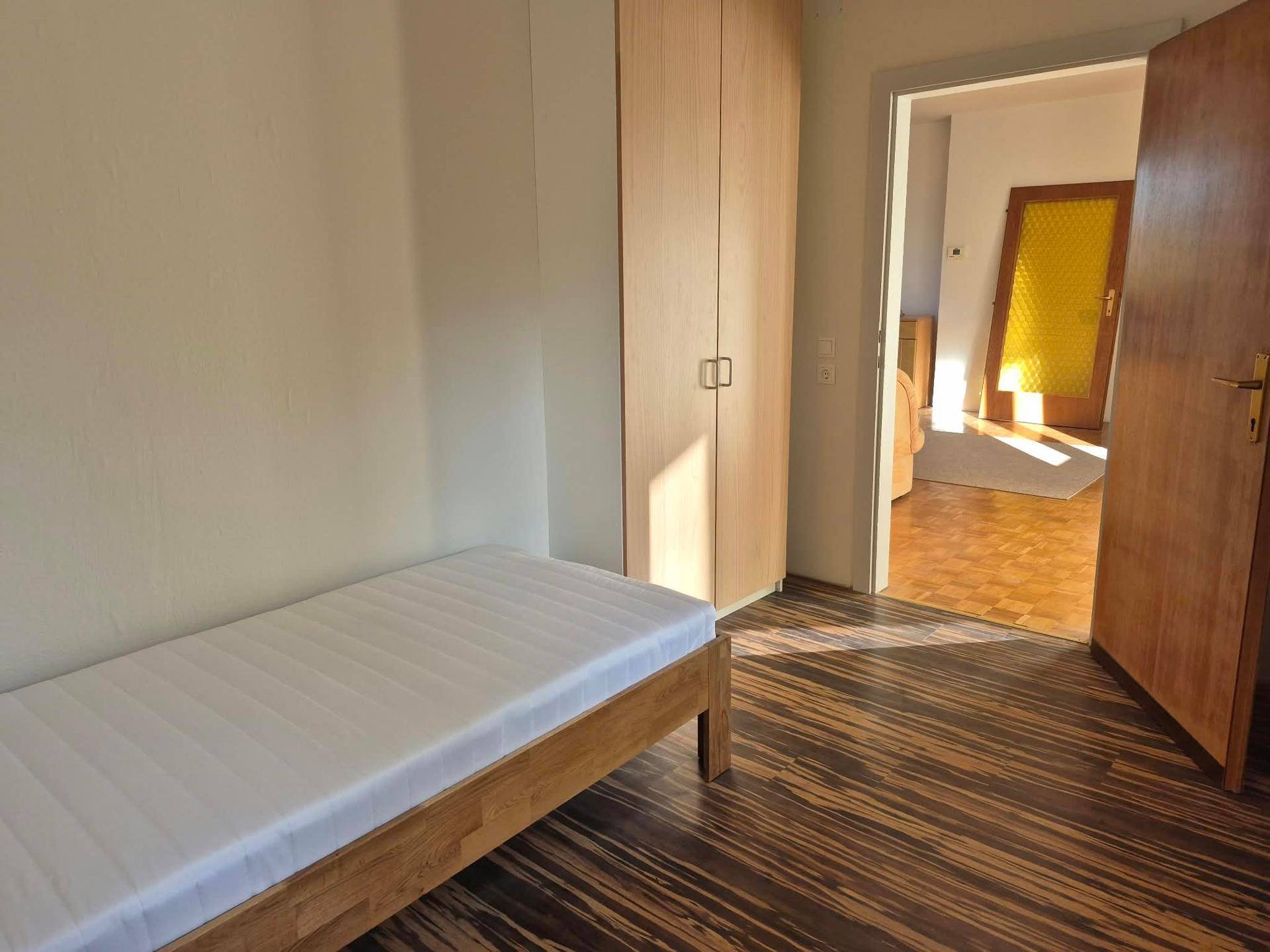 Schlafzimmer mit Einzelbett, Kleiderschrank und dunklem Holzboden, durchflutet von Sonnenlicht.