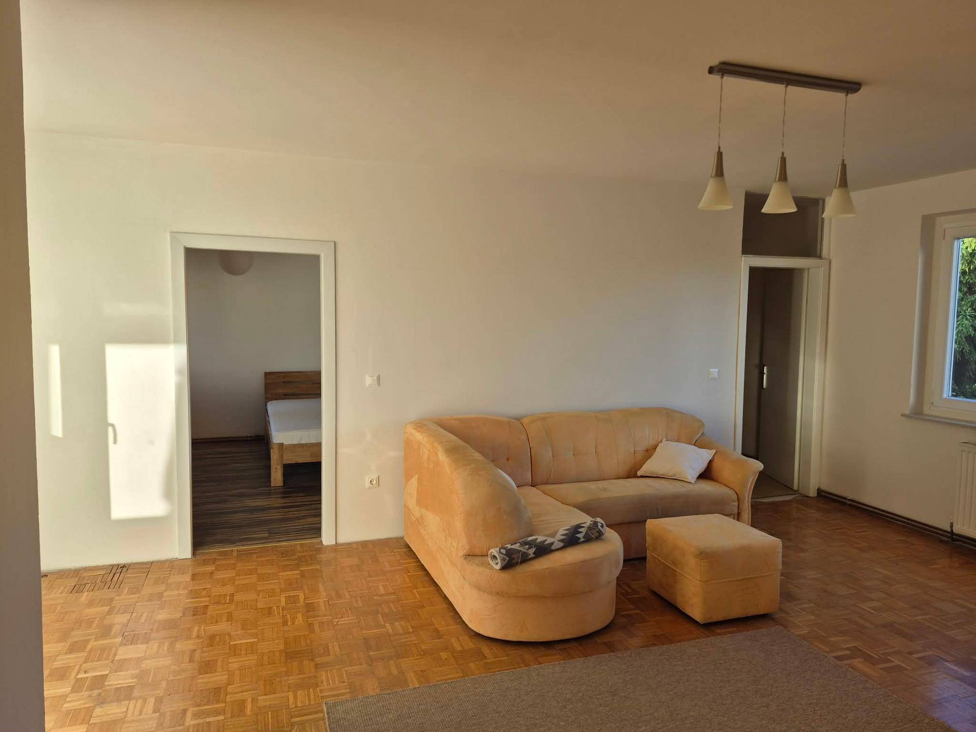 Gemütliches Wohnzimmer mit beigem Sofa, Parkettboden und einem offenen Durchgang zu einem Schlafzimmer.