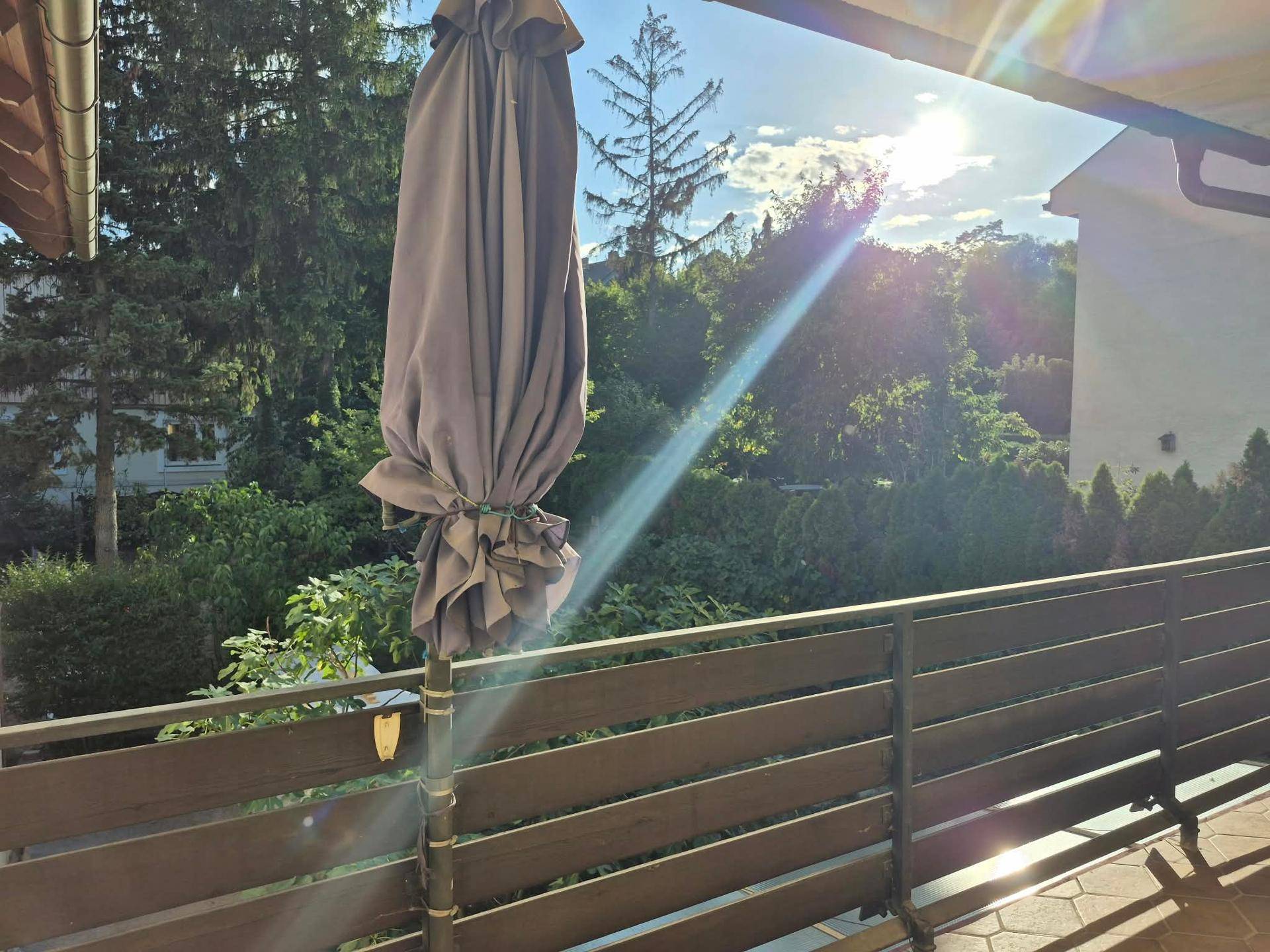 Sonniger Balkon mit großem Sonnenschirm und Blick auf üppiges Grün und Bäume.